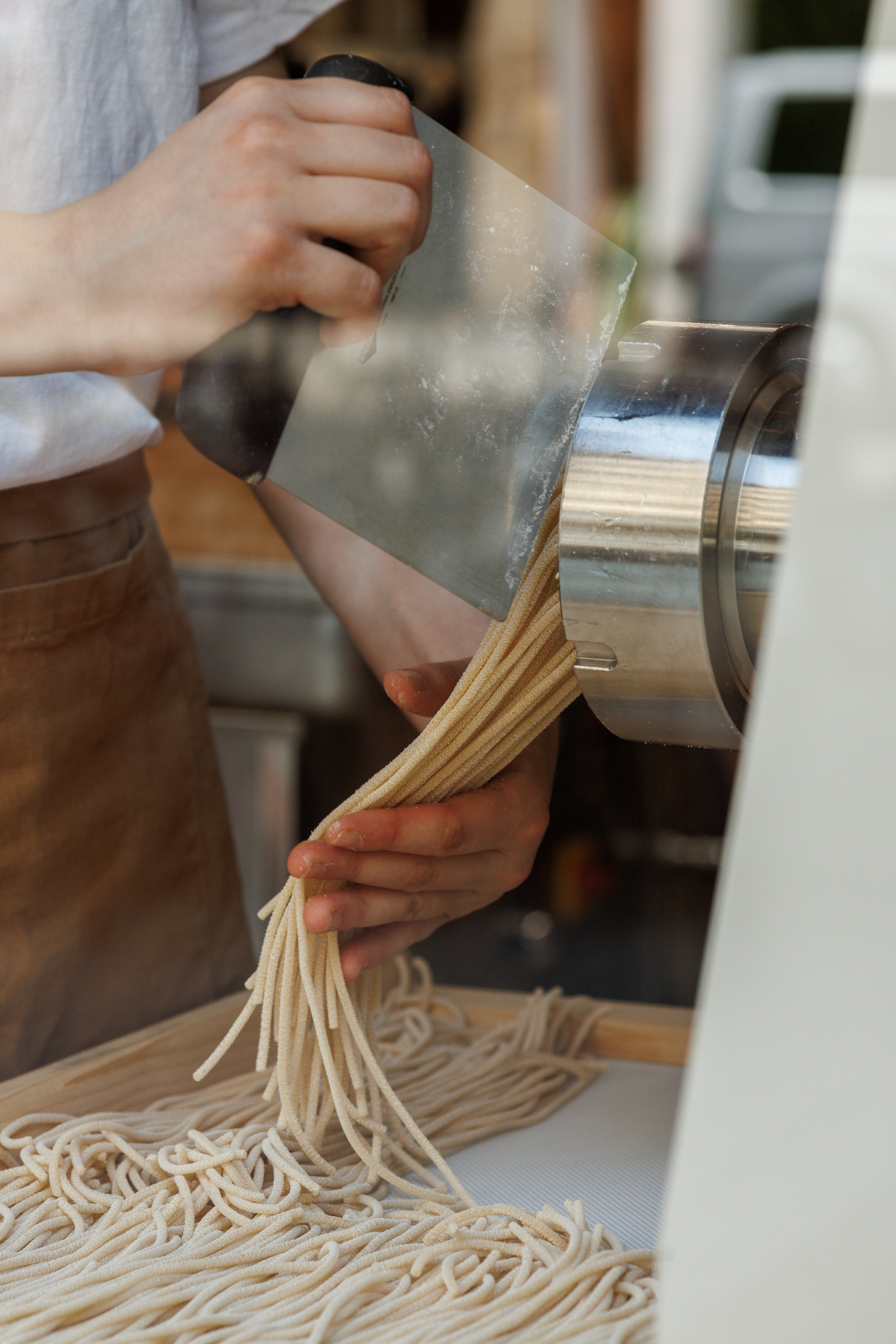 A person uses a metal scraper to cut fresh pasta strands as they emerge from an industrial machine.