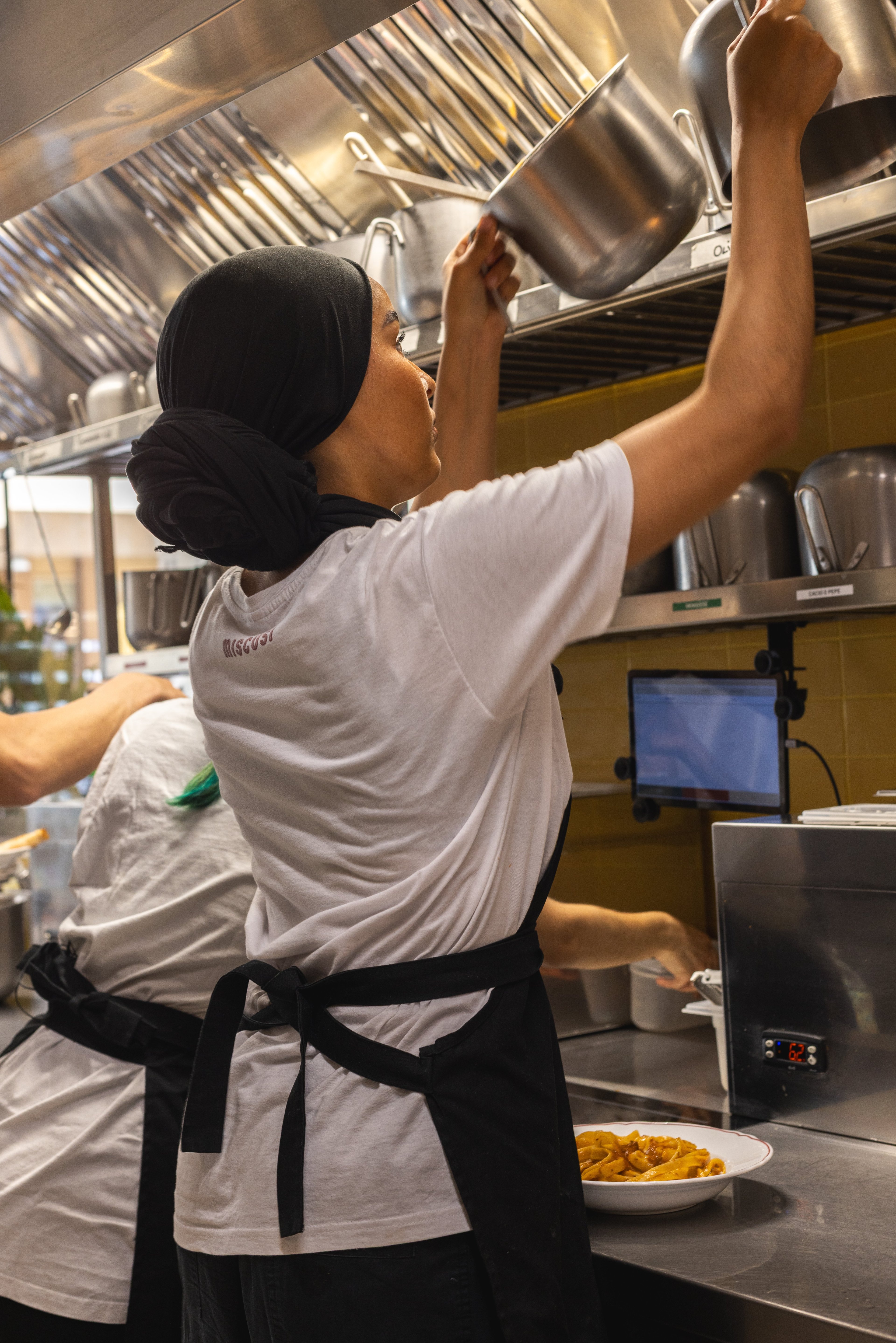 A chef in a professional kitchen prepares a pasta dish while working under bright, functional lighting.