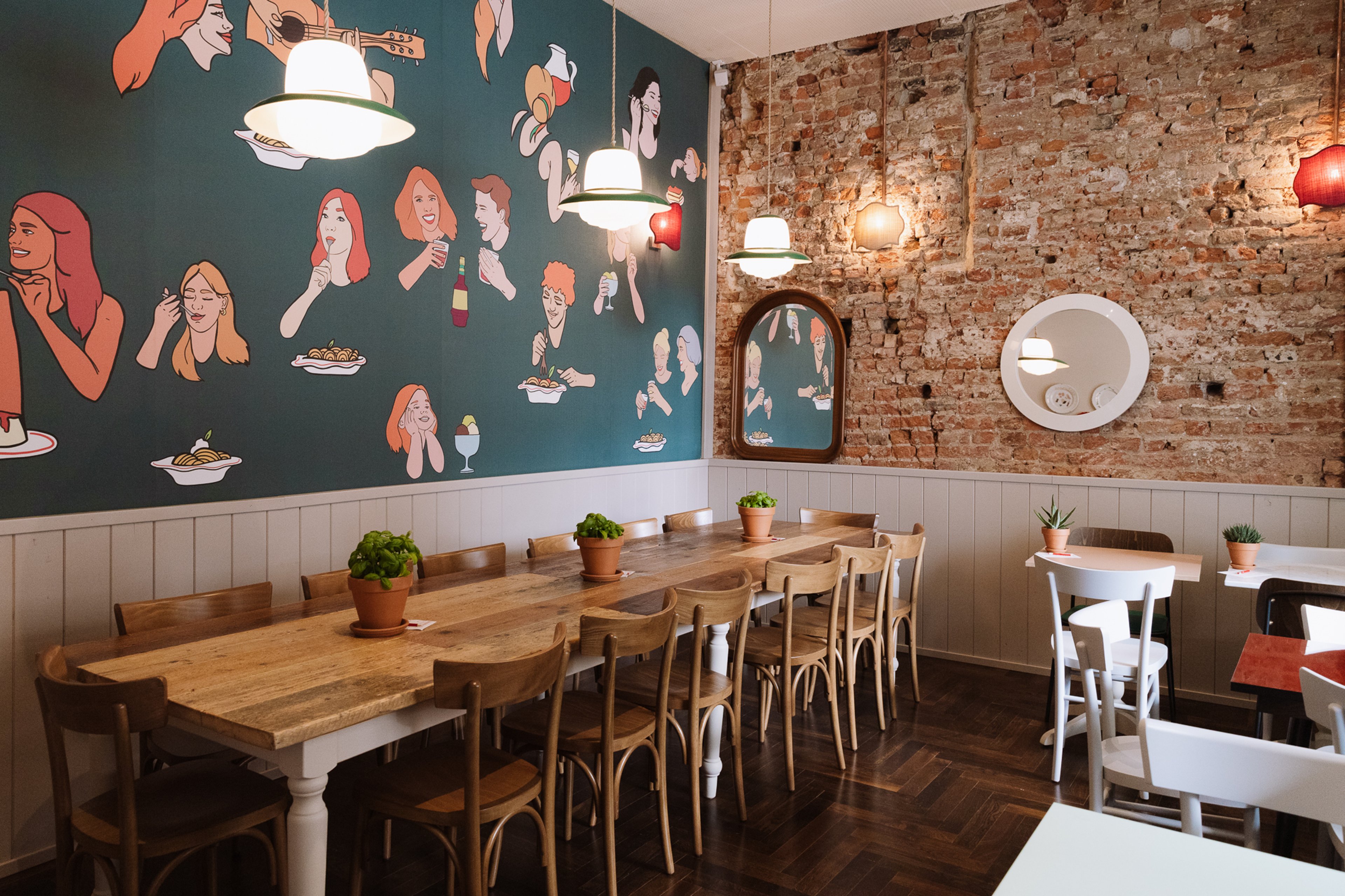 A cozy restaurant interior features a long wooden table, vibrant illustrated wallpaper, and warm pendant lighting against an exposed brick wall.