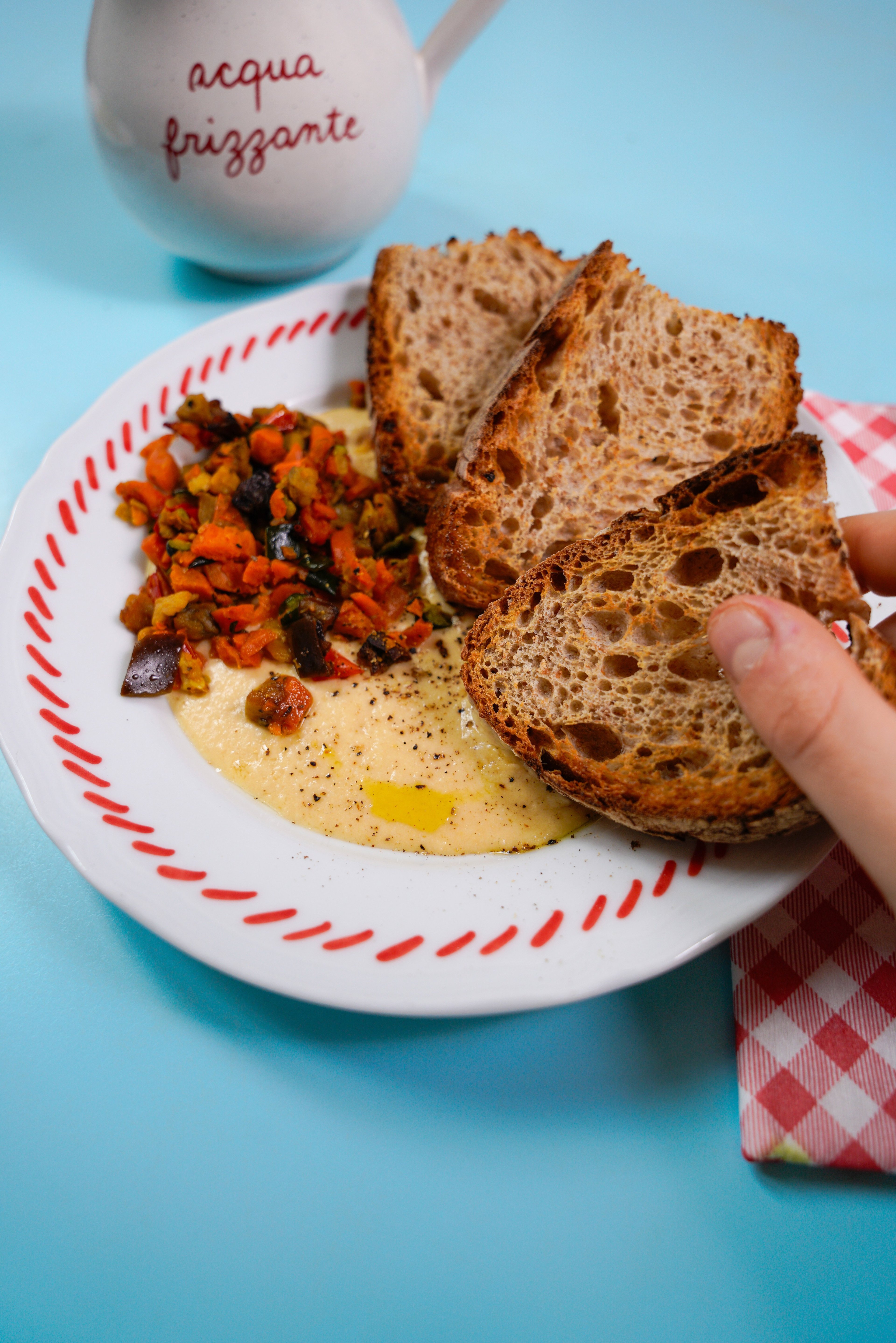 A hand holds a slice of toasted sourdough bread over a plate of creamy hummus topped with roasted vegetables.