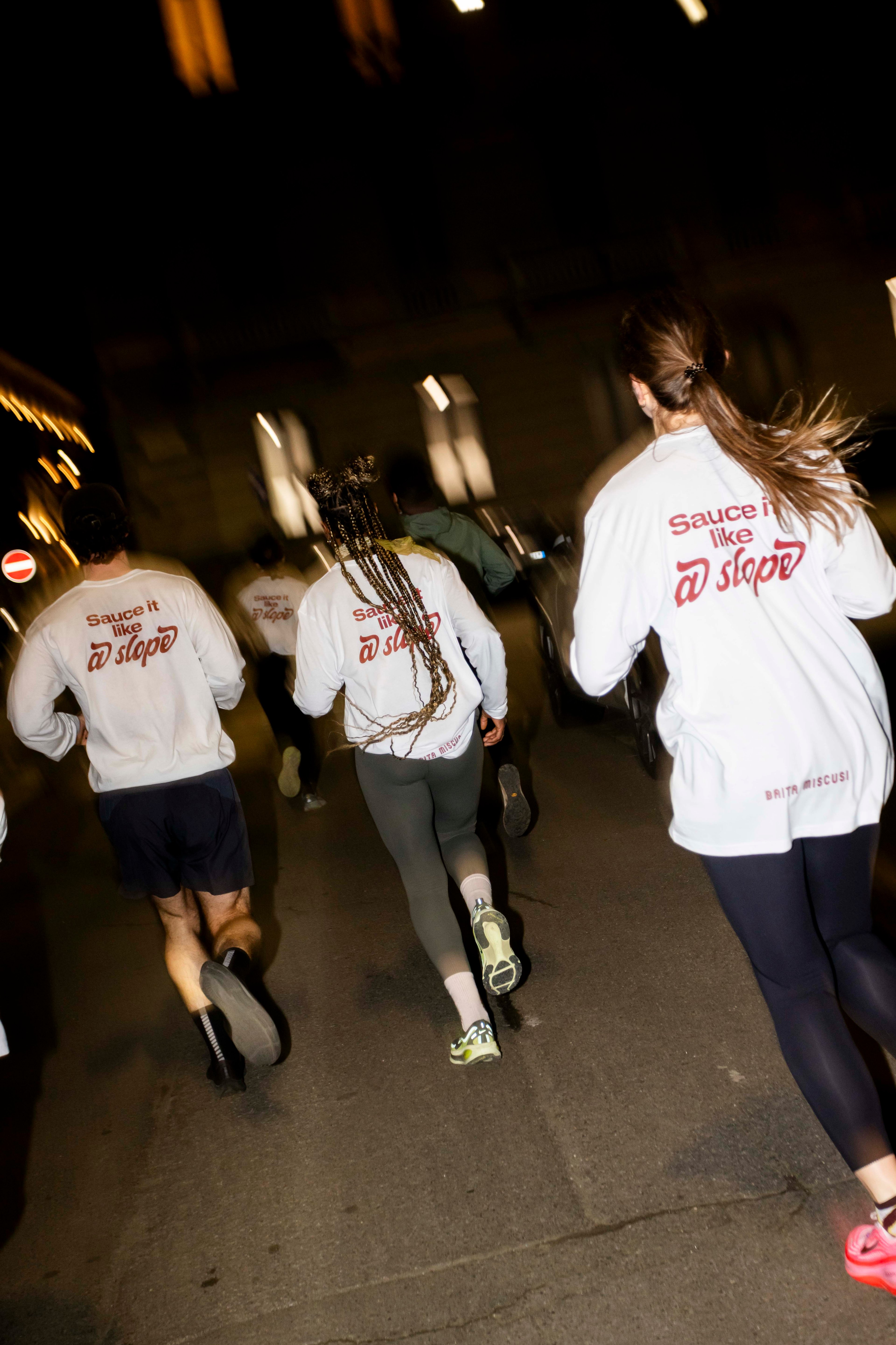 A group of people wearing branded long-sleeve shirts run together on a city street at night.