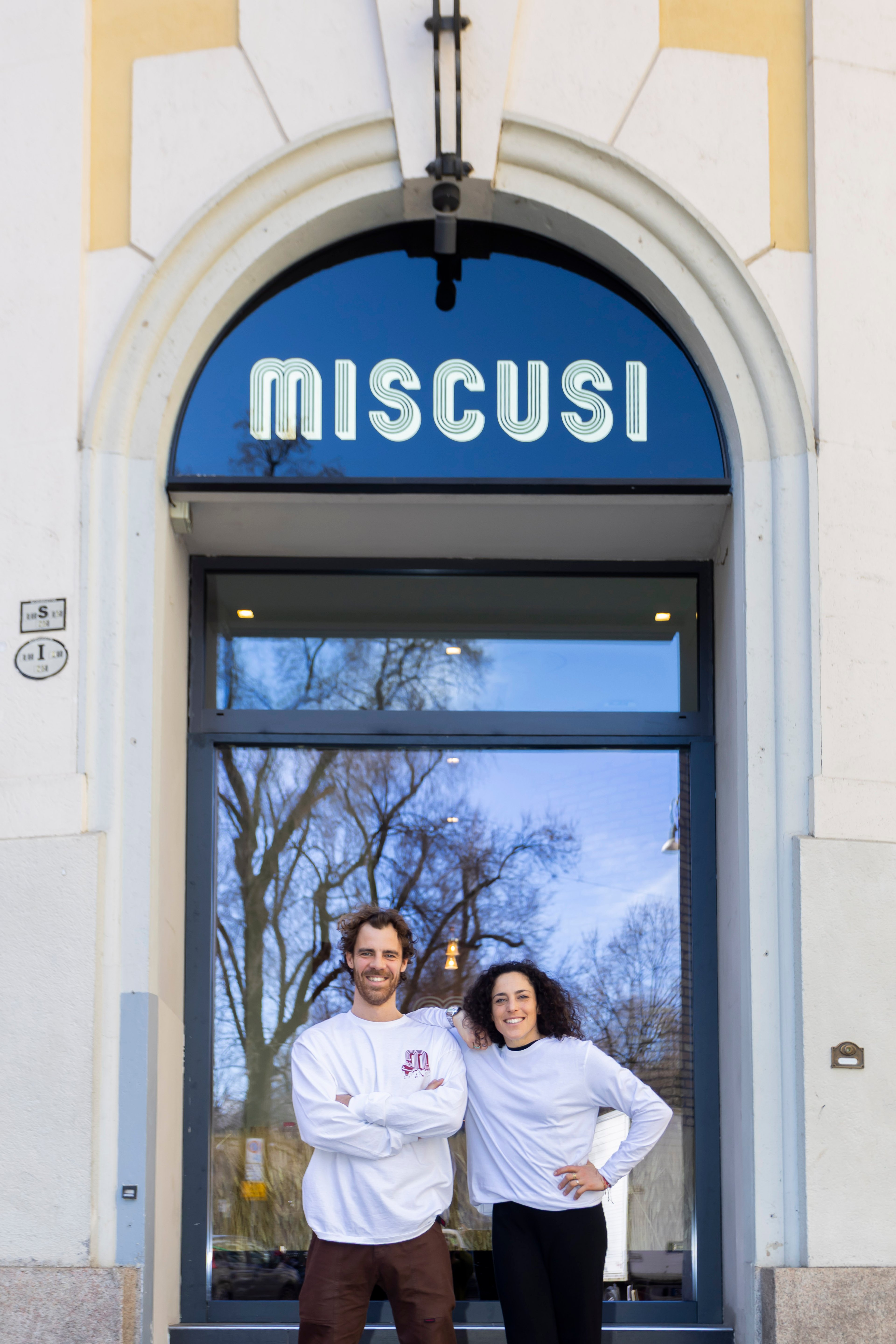 A man and a woman stand smiling in front of the entrance to a restaurant named Miscusi.