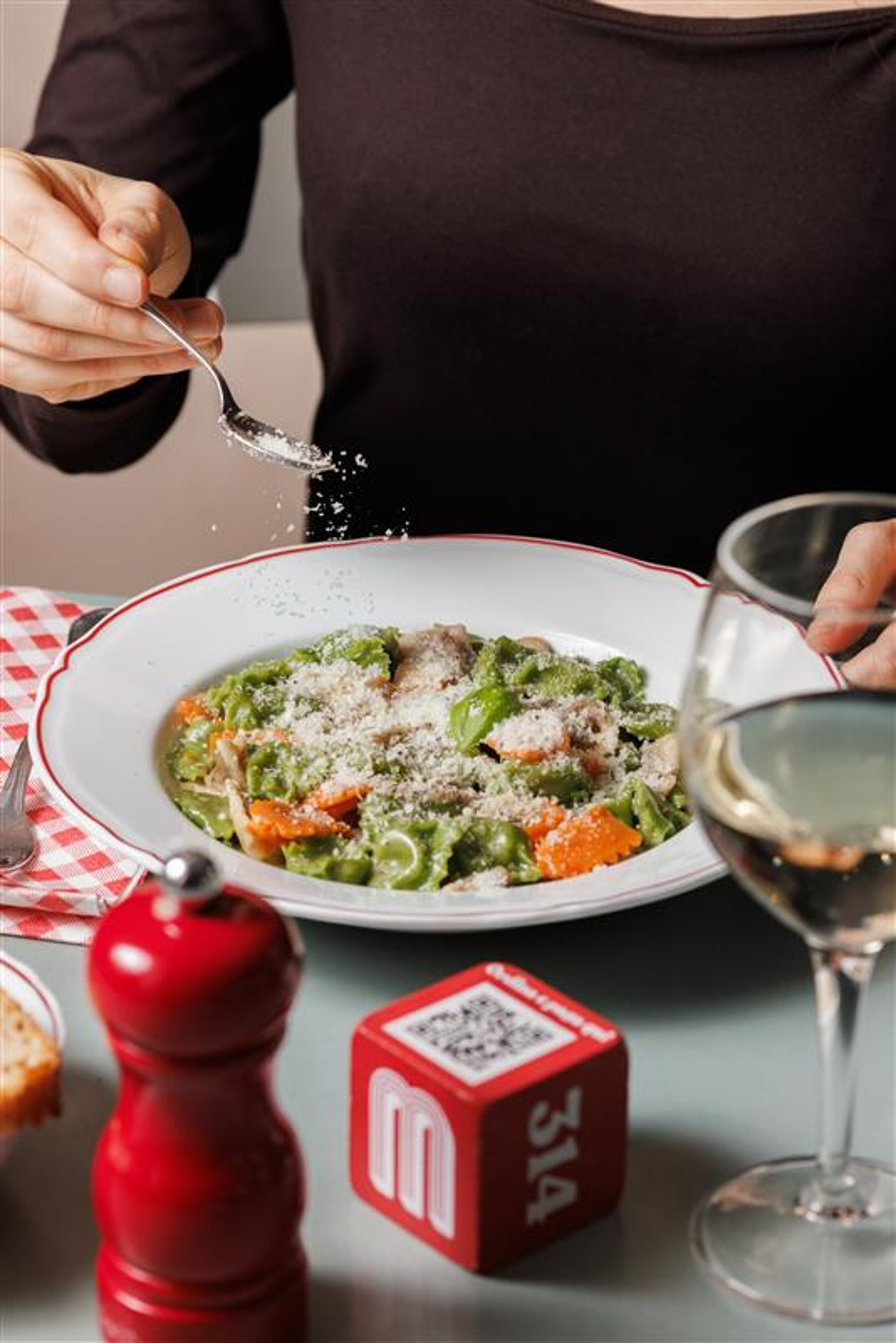 A person sprinkles grated cheese over a vibrant plate of green and orange pasta in a cozy restaurant setting.