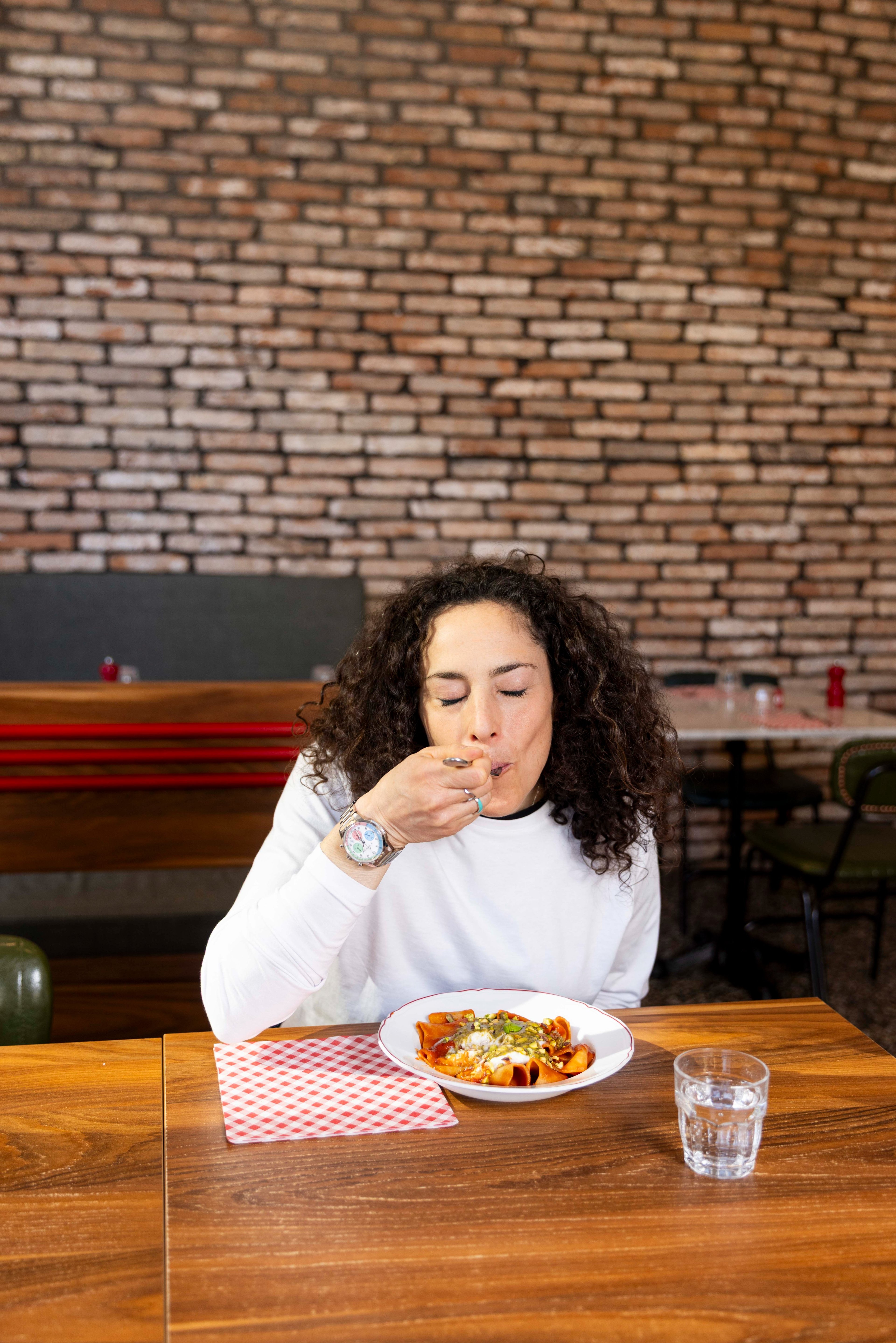 A woman with curly hair enjoys a plate of pasta in a restaurant with a rustic brick wall background.