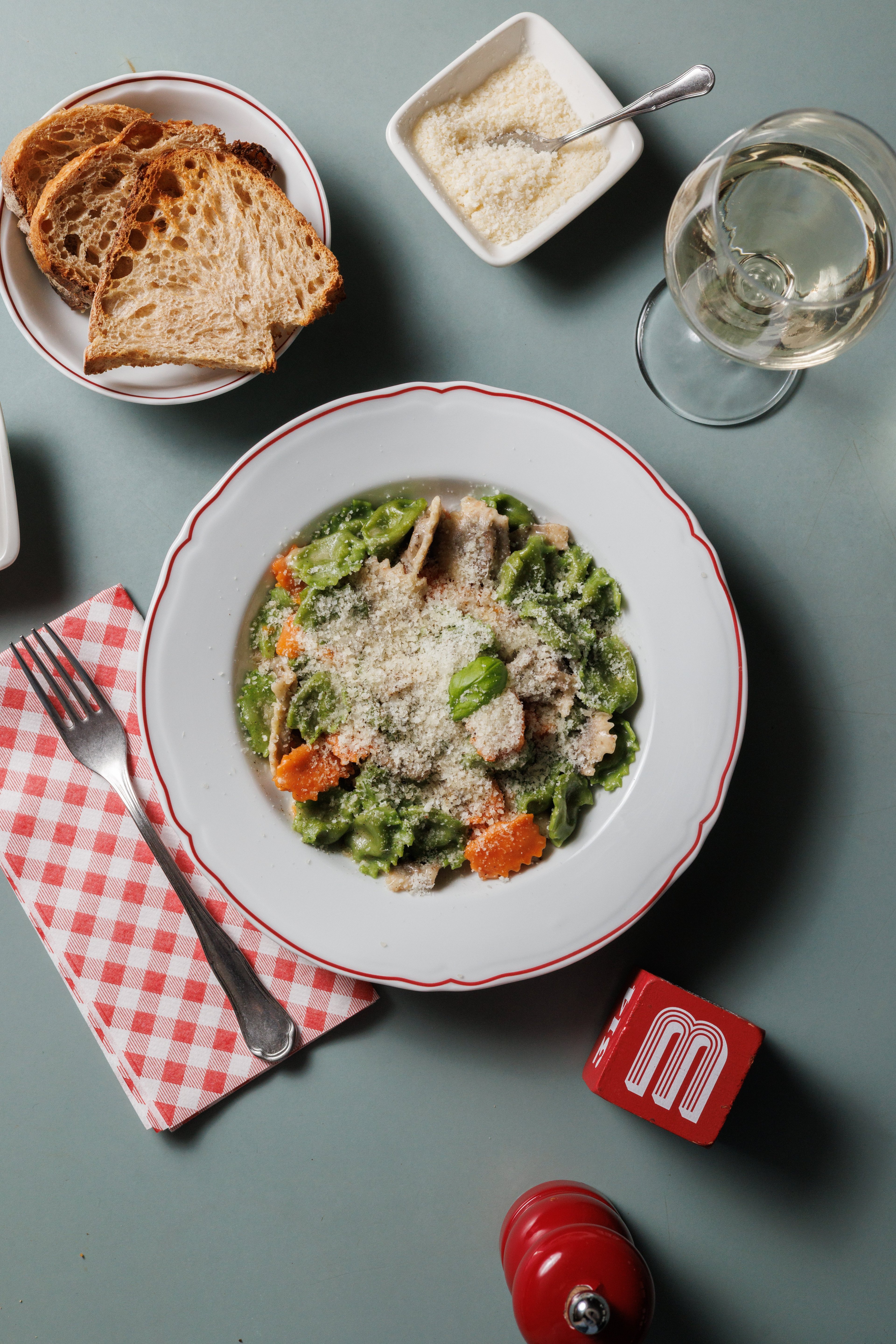 A top-down view of a plate of colorful pasta served with toasted bread, grated cheese, and a glass of white wine.