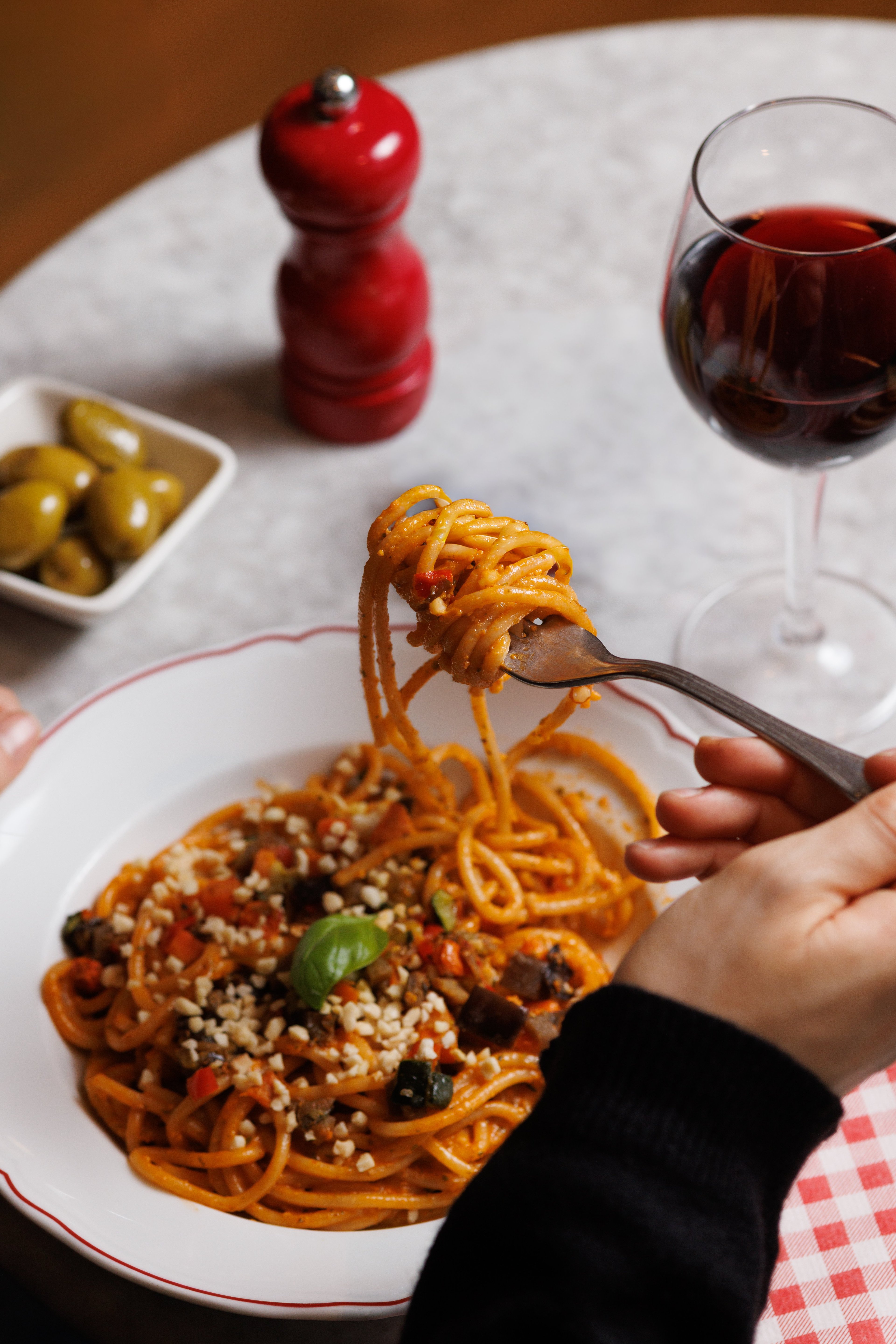 A person enjoys a plate of pasta with vegetables and nuts, accompanied by a glass of red wine.