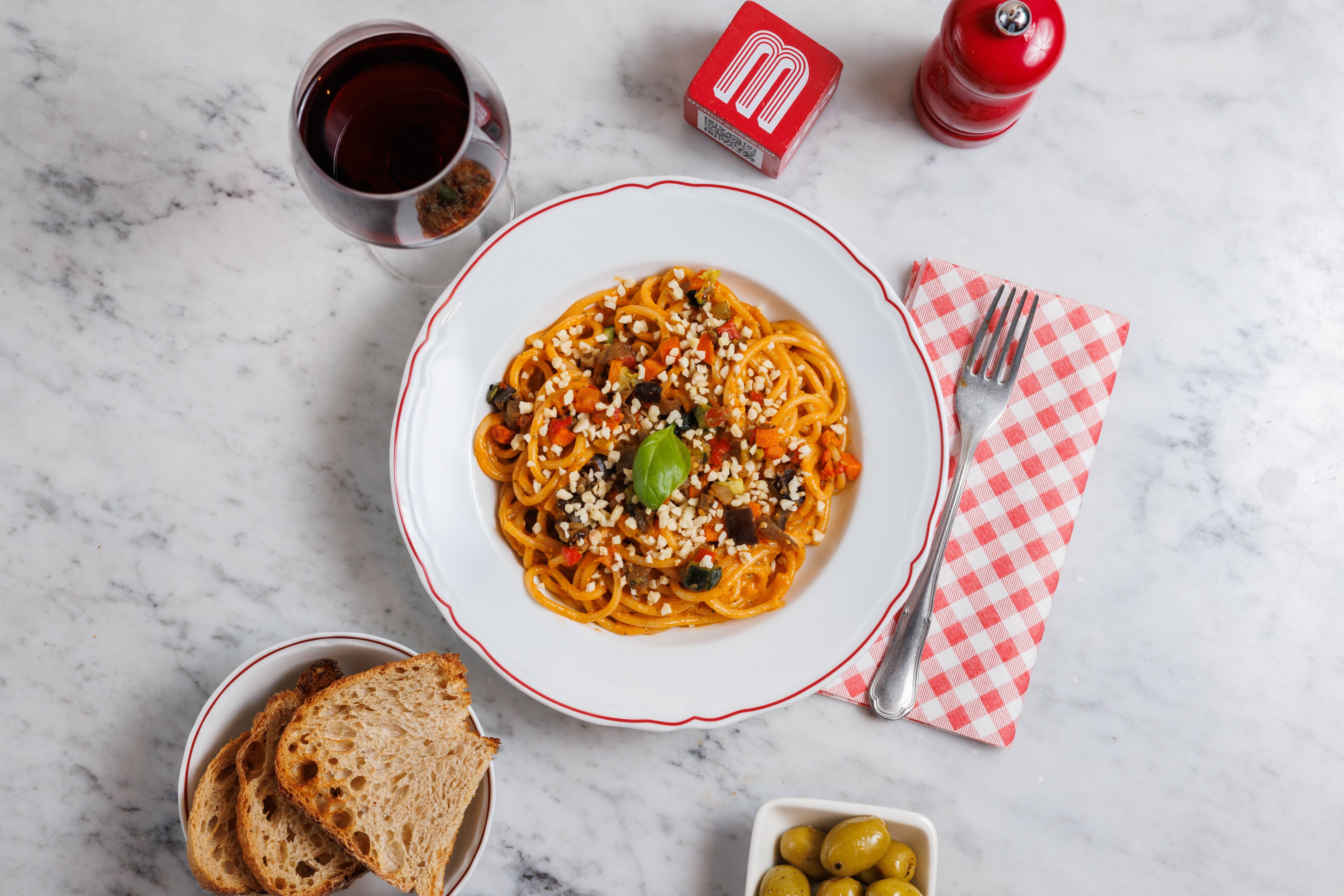 A top-down view of a plate of pasta with vegetables and nuts, served with bread, olives, and red wine on a marble table.