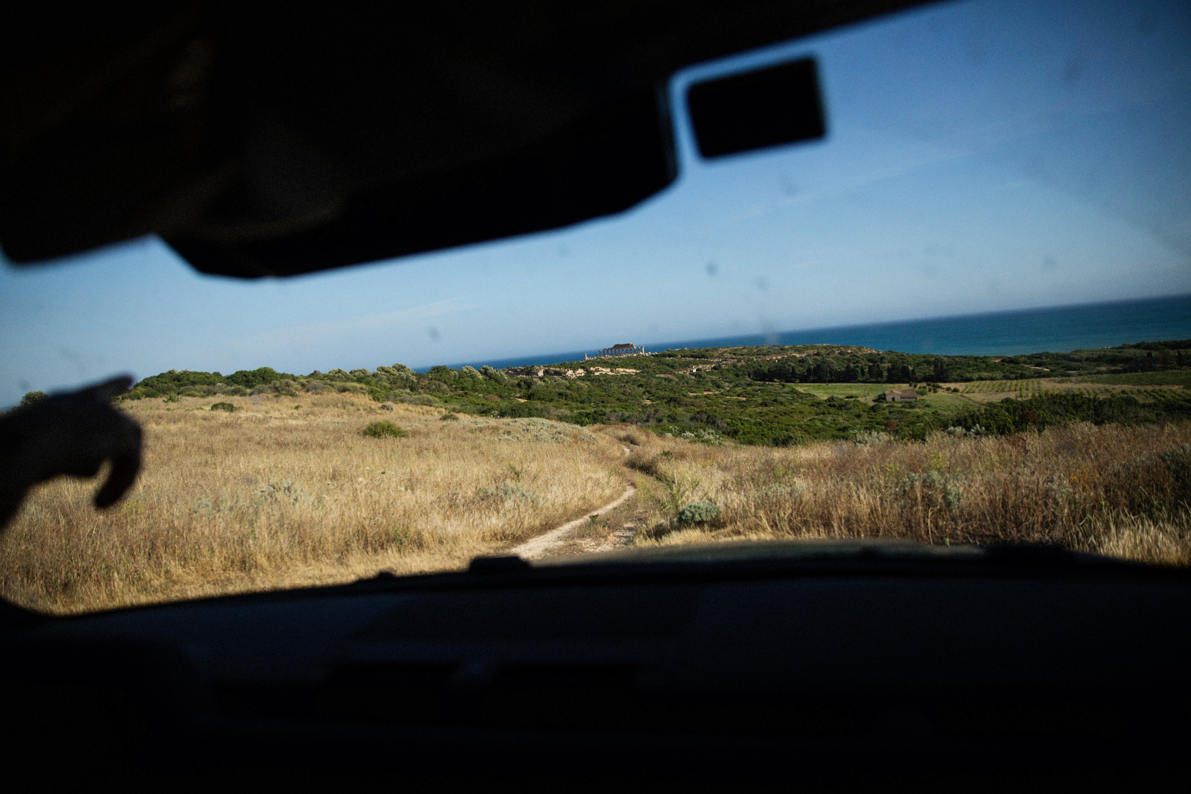 A view from inside a vehicle looking out over a grassy landscape toward a distant ancient temple by the sea.