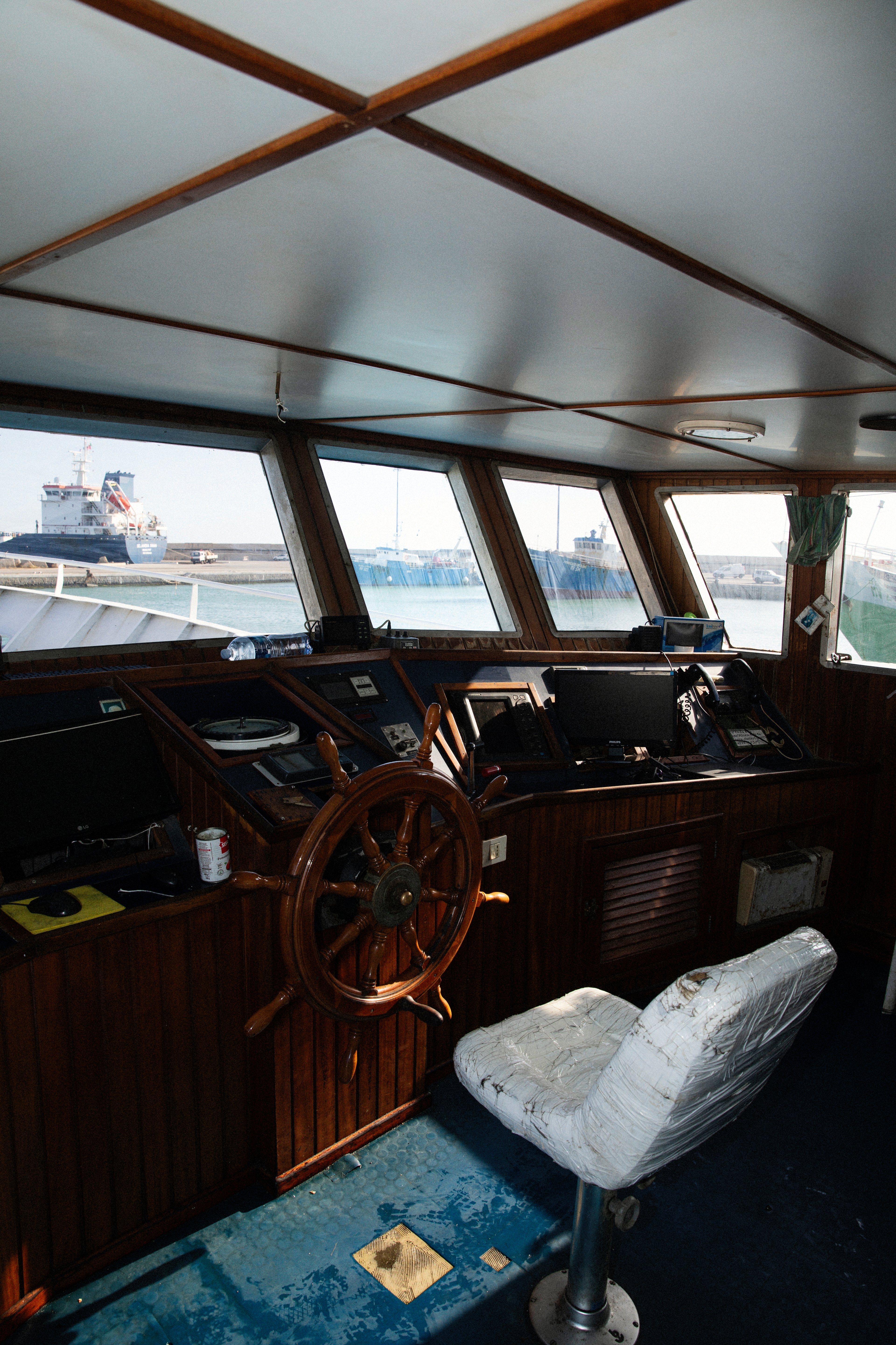 The interior of a ship's bridge features a wooden steering wheel and a worn chair overlooking a harbor.