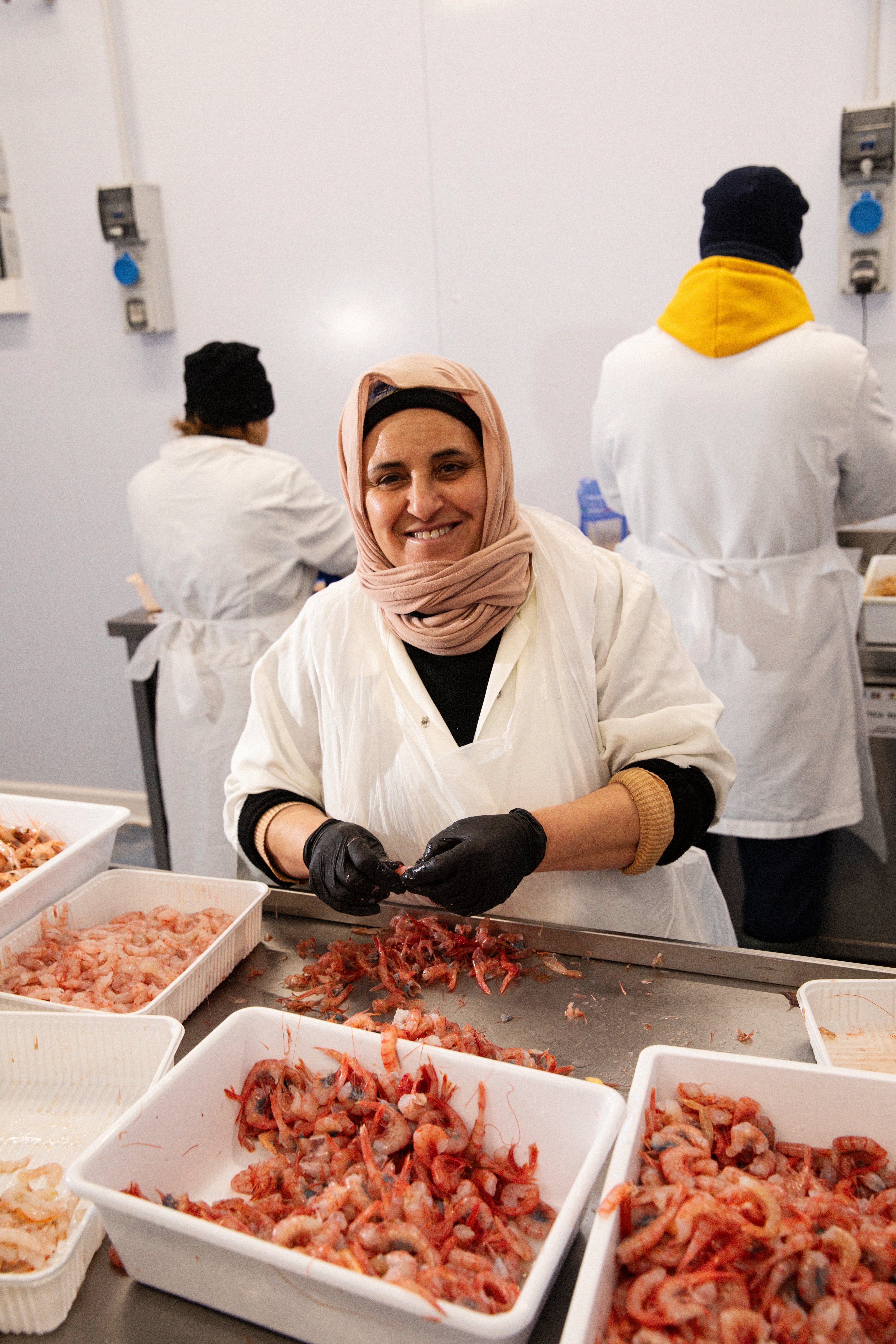 A smiling woman in a headscarf and apron processes fresh shrimp in a brightly lit industrial kitchen.