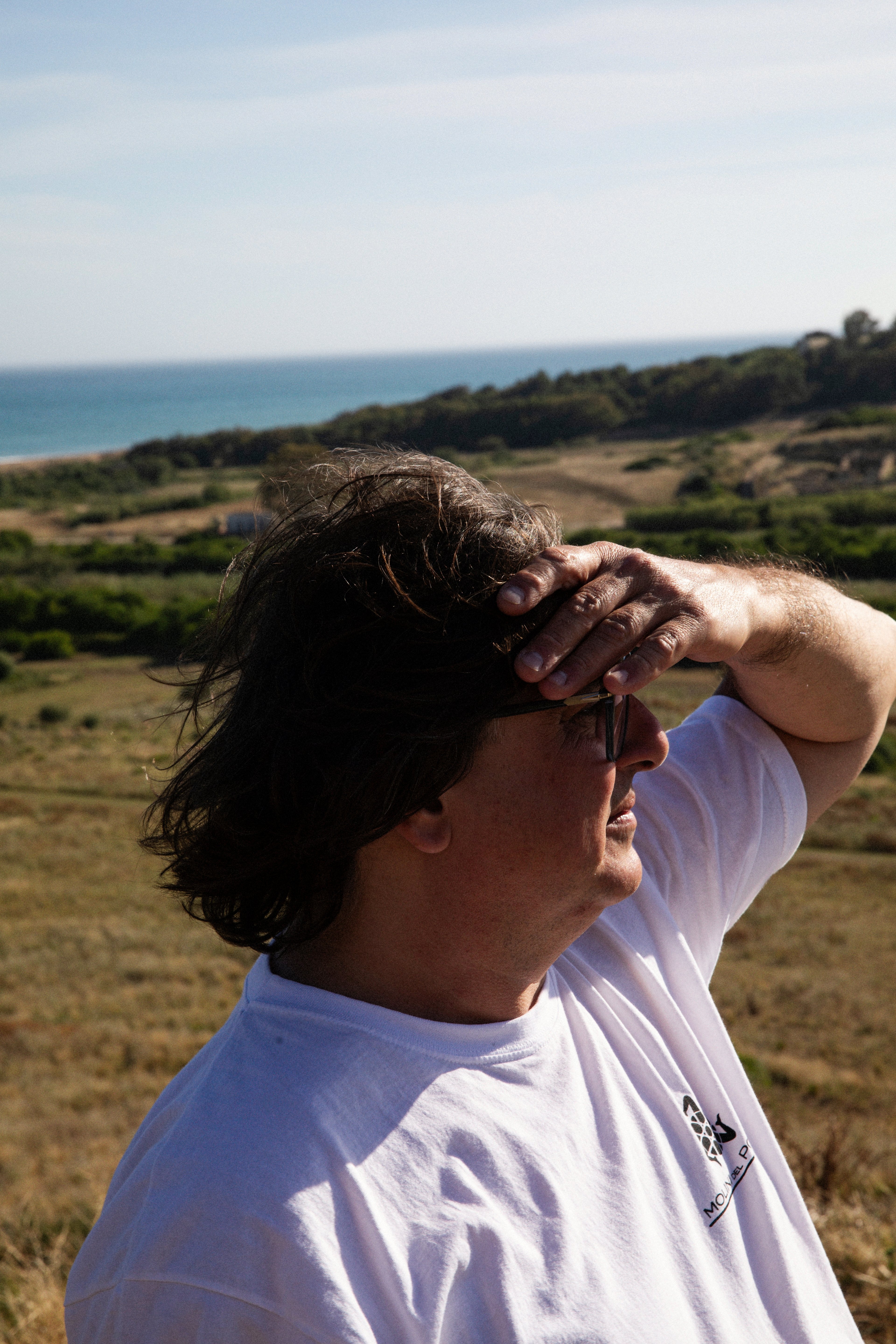 A man stands outdoors in a sunny, natural landscape, shielding his eyes from the bright light.