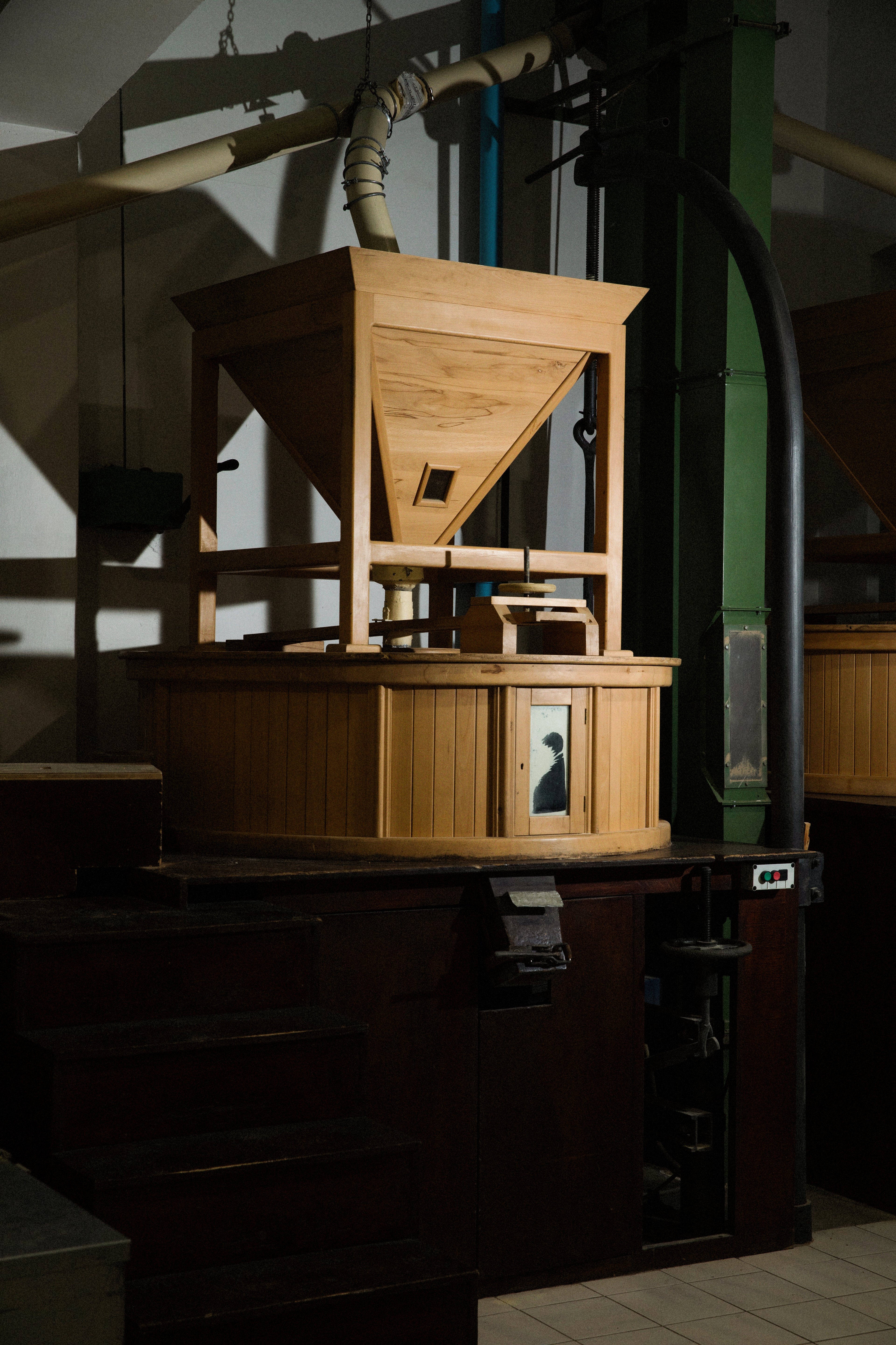 A vintage wooden grain mill stands in a dimly lit, industrial-style room.