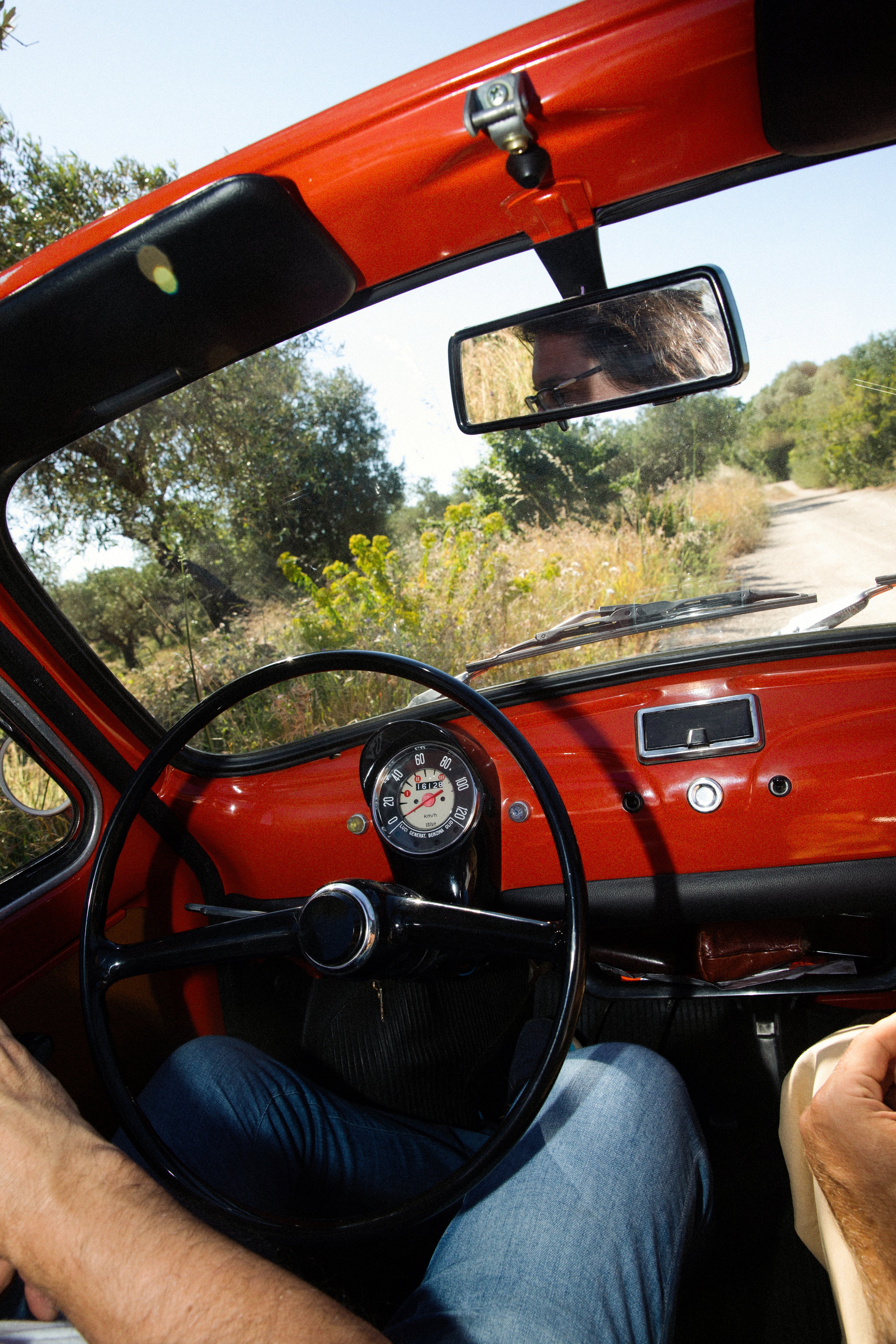 A driver's perspective from inside a vintage red car traveling along a sunny, tree-lined country road.