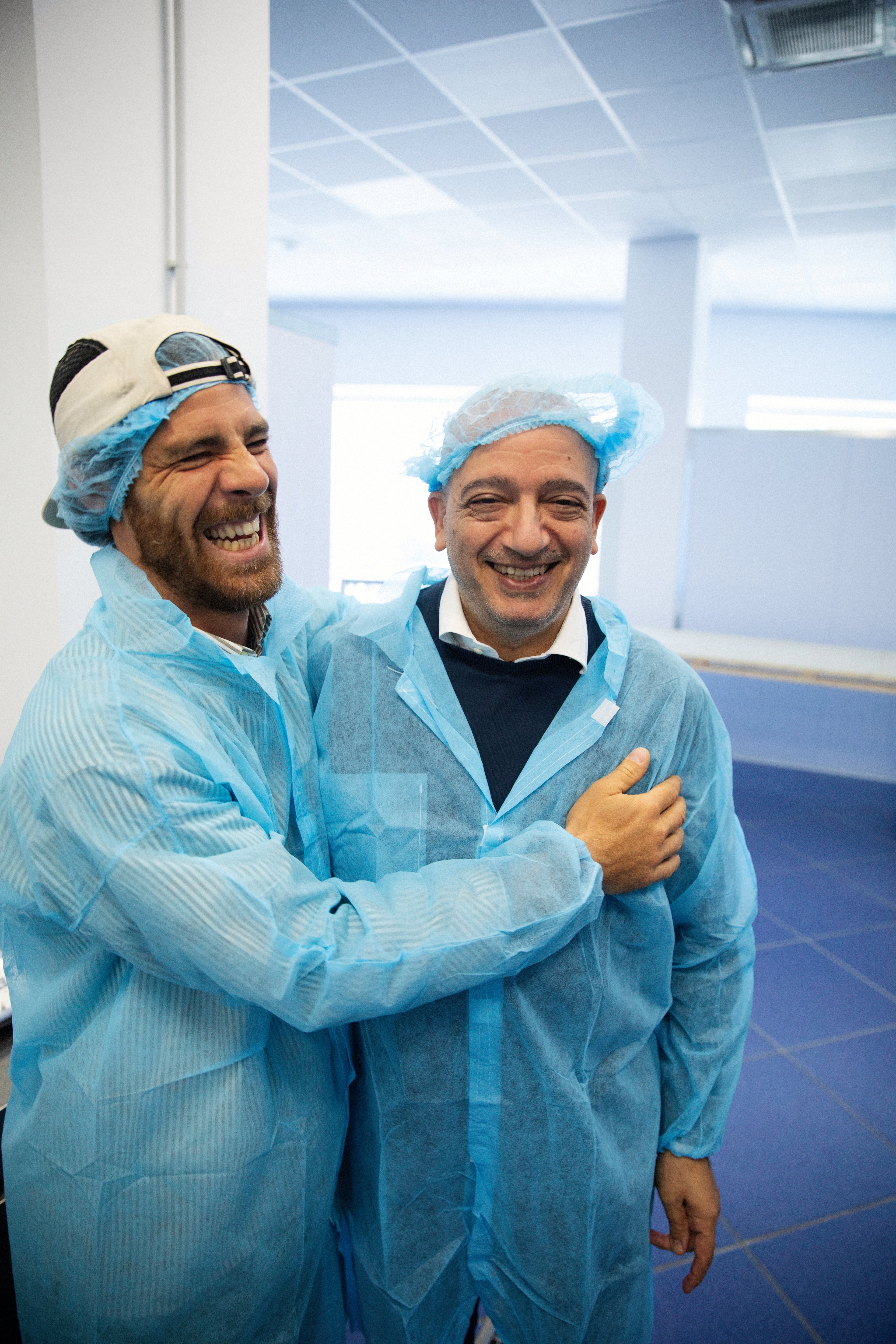 Two men wearing protective medical gear and hairnets laugh together in a bright, clinical environment.