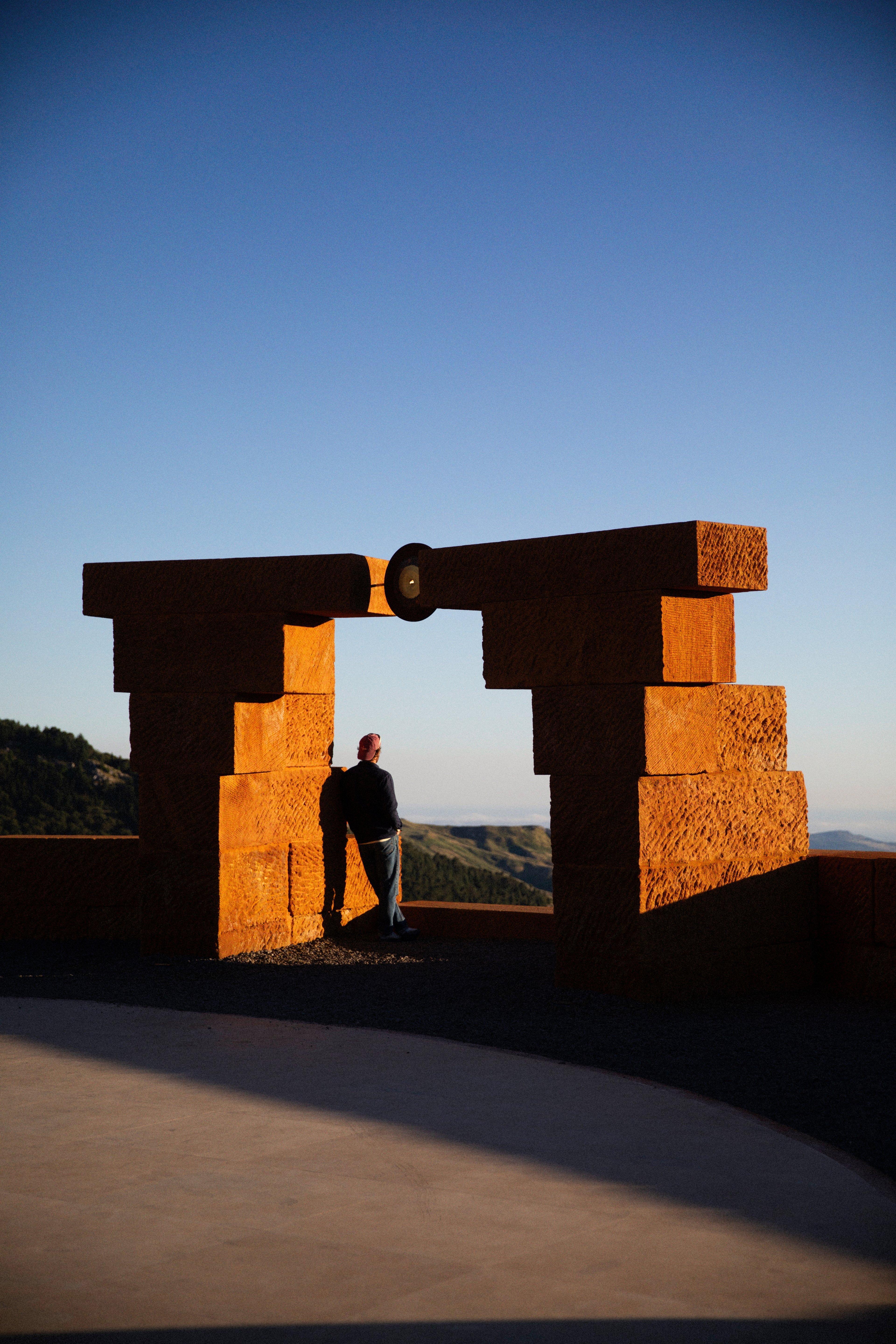 A person stands leaning against a large, geometric stone sculpture overlooking a mountainous landscape at sunset.