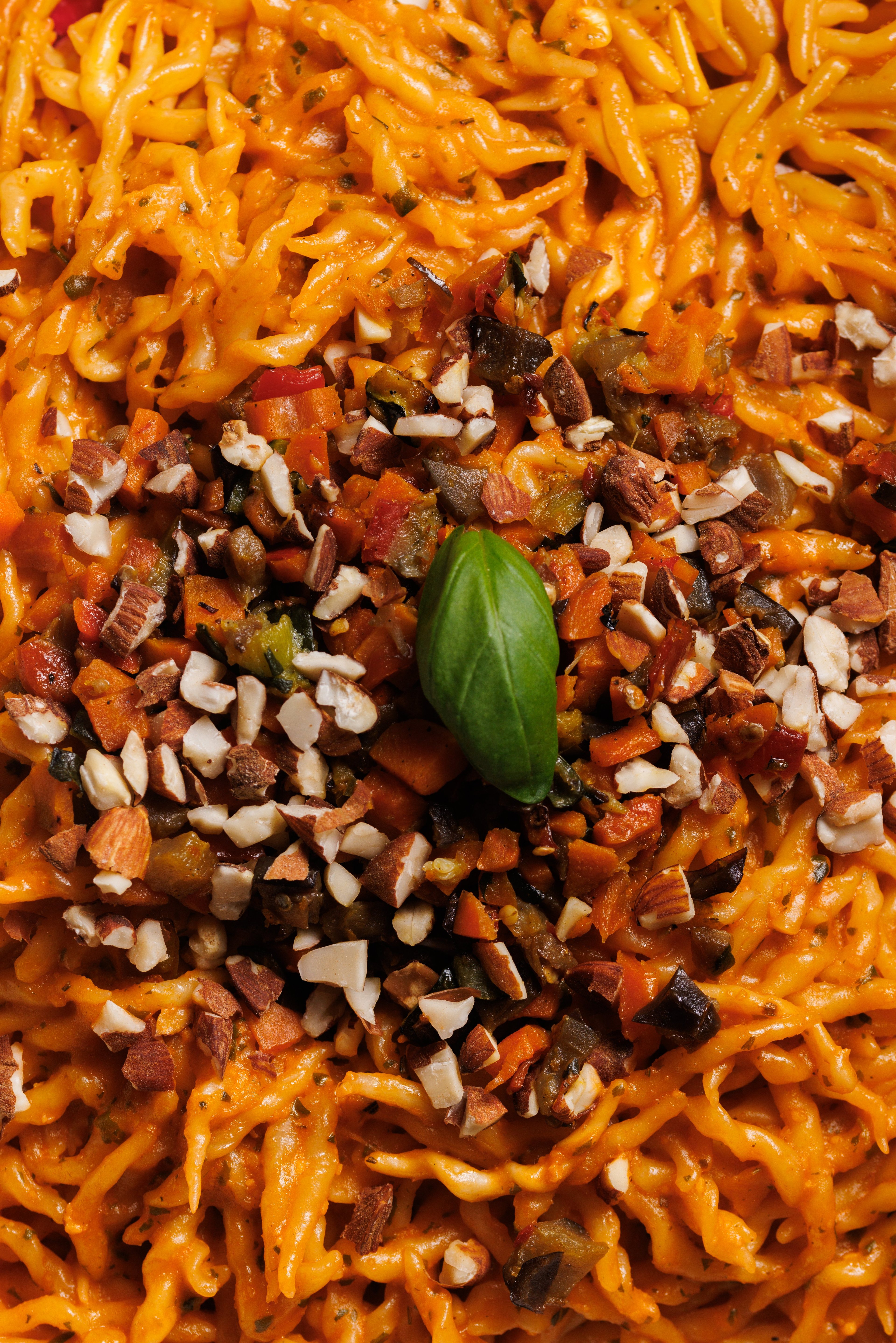 A top-down view of a vibrant pasta dish topped with chopped nuts, diced vegetables, and a fresh basil leaf.