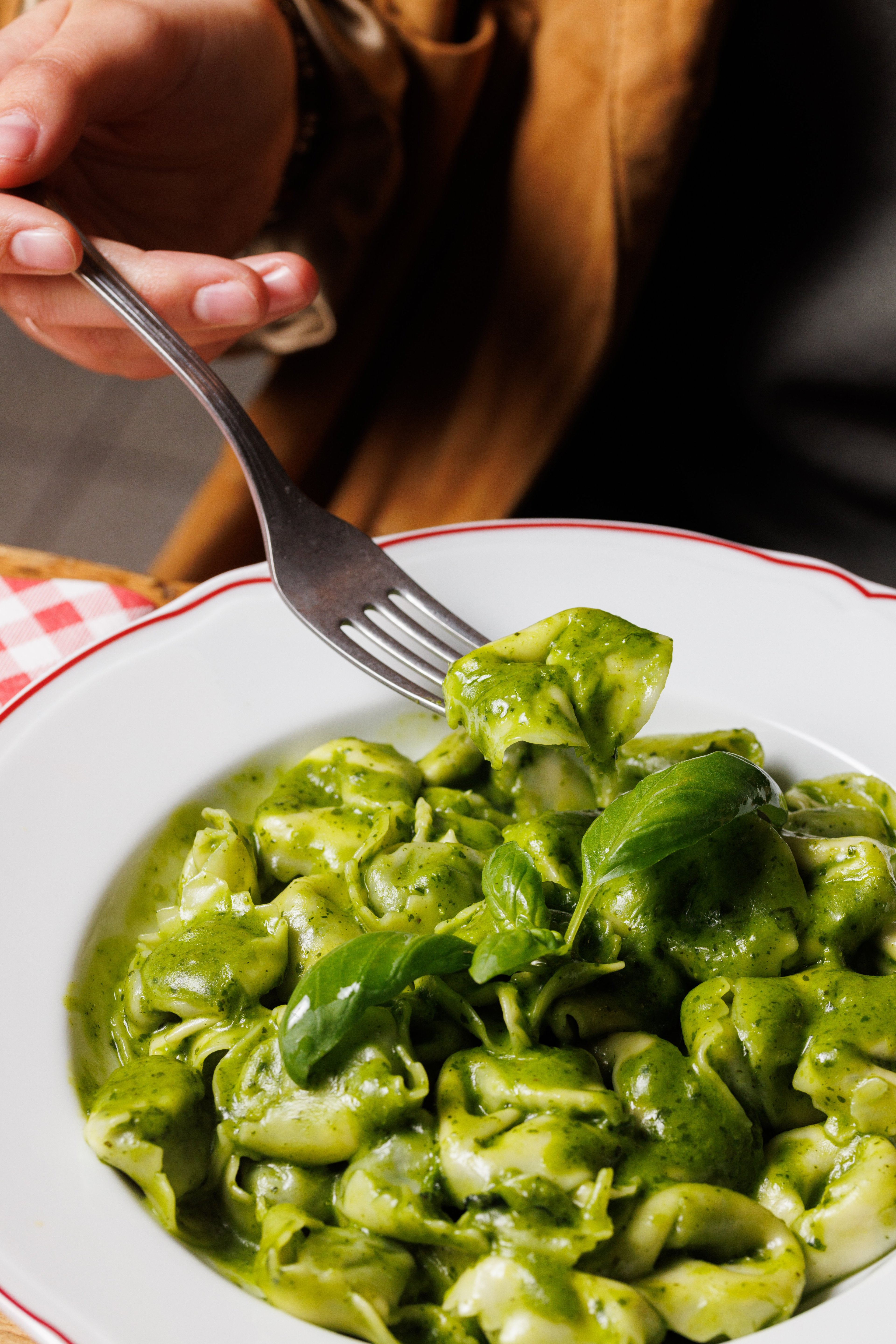 A person uses a fork to lift a piece of tortellini coated in vibrant green pesto sauce from a white plate.