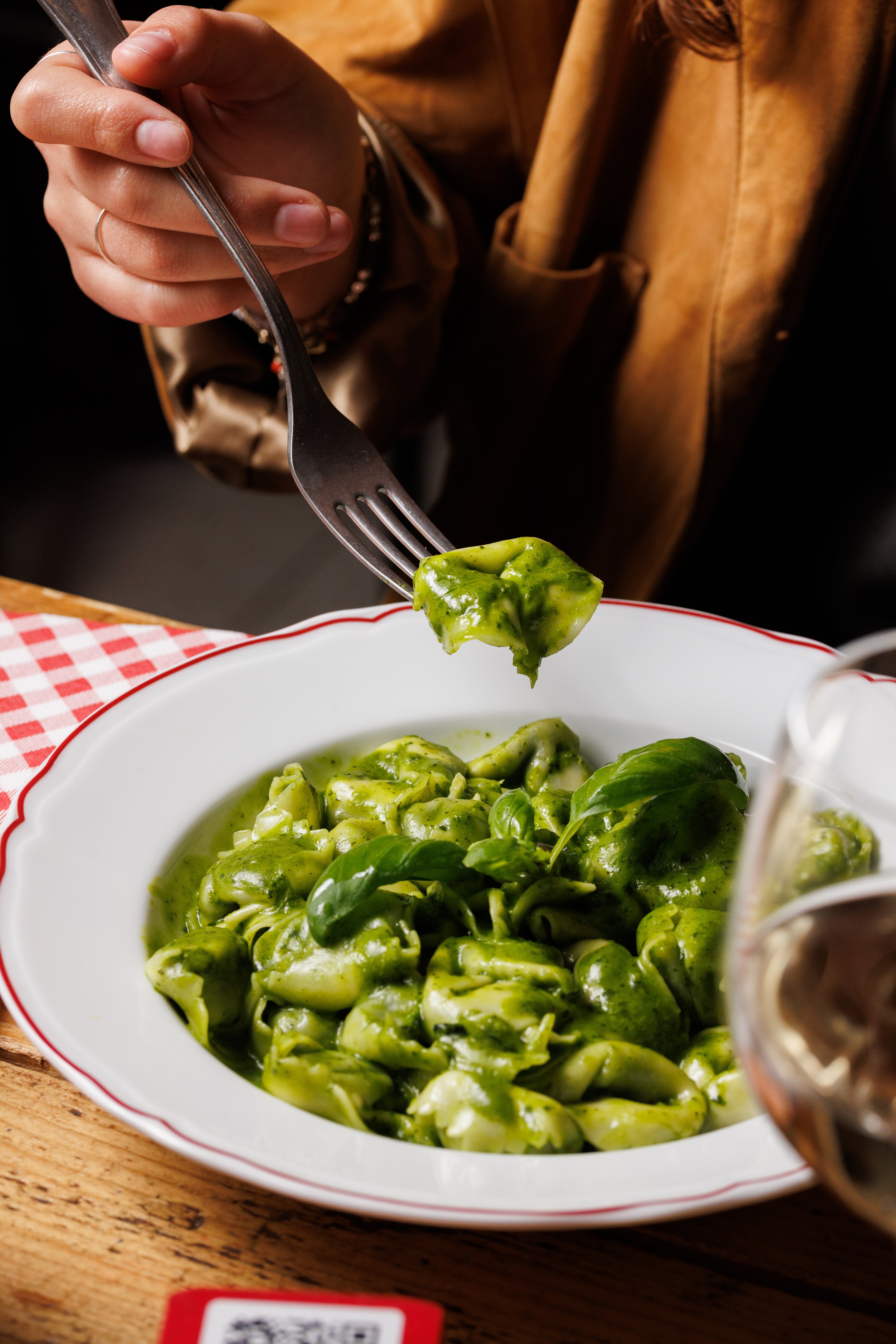 A person uses a fork to lift a piece of tortellini coated in vibrant green pesto sauce from a white plate.