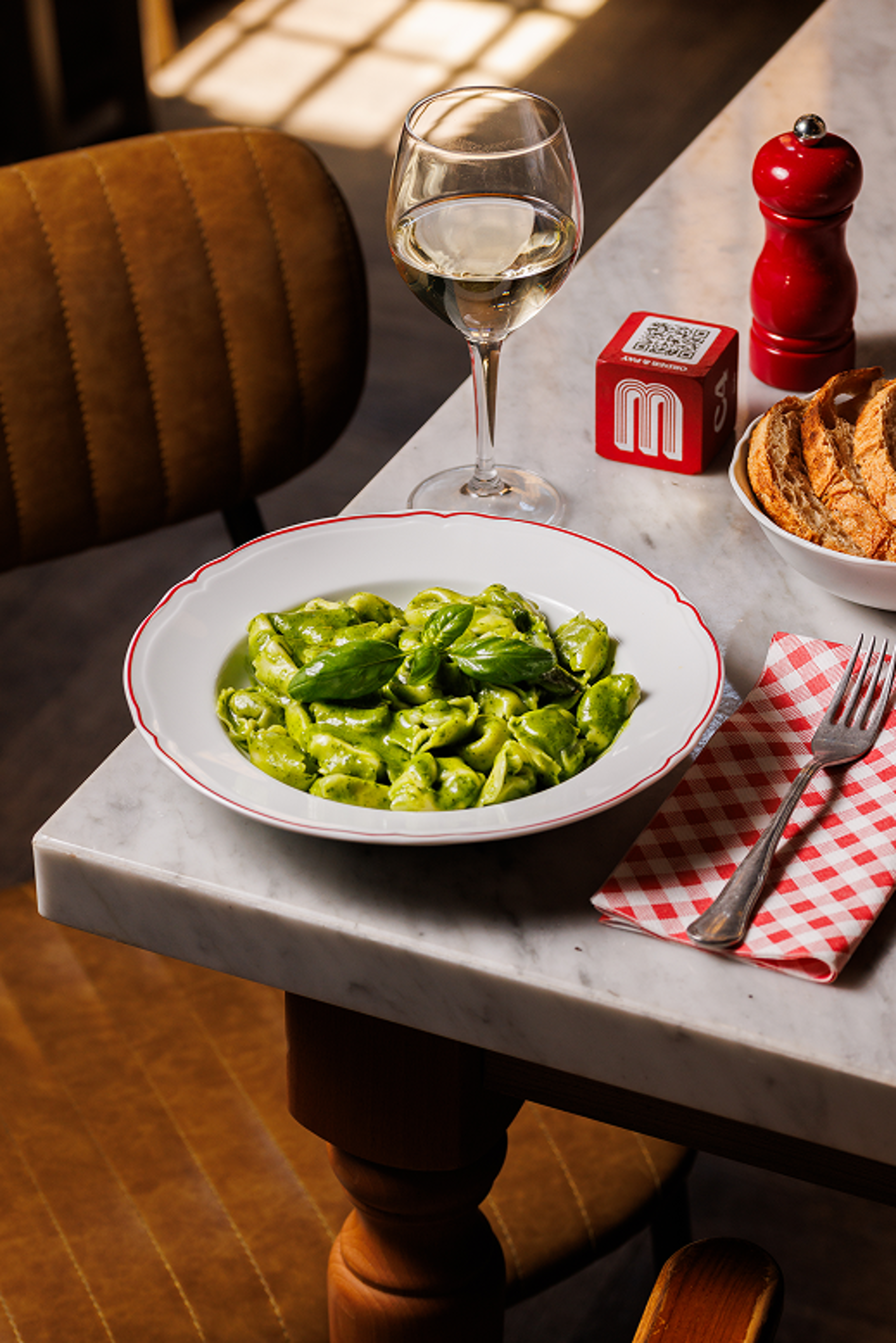 A plate of fresh green pesto tortellini is served on a marble table alongside a glass of white wine and rustic bread.