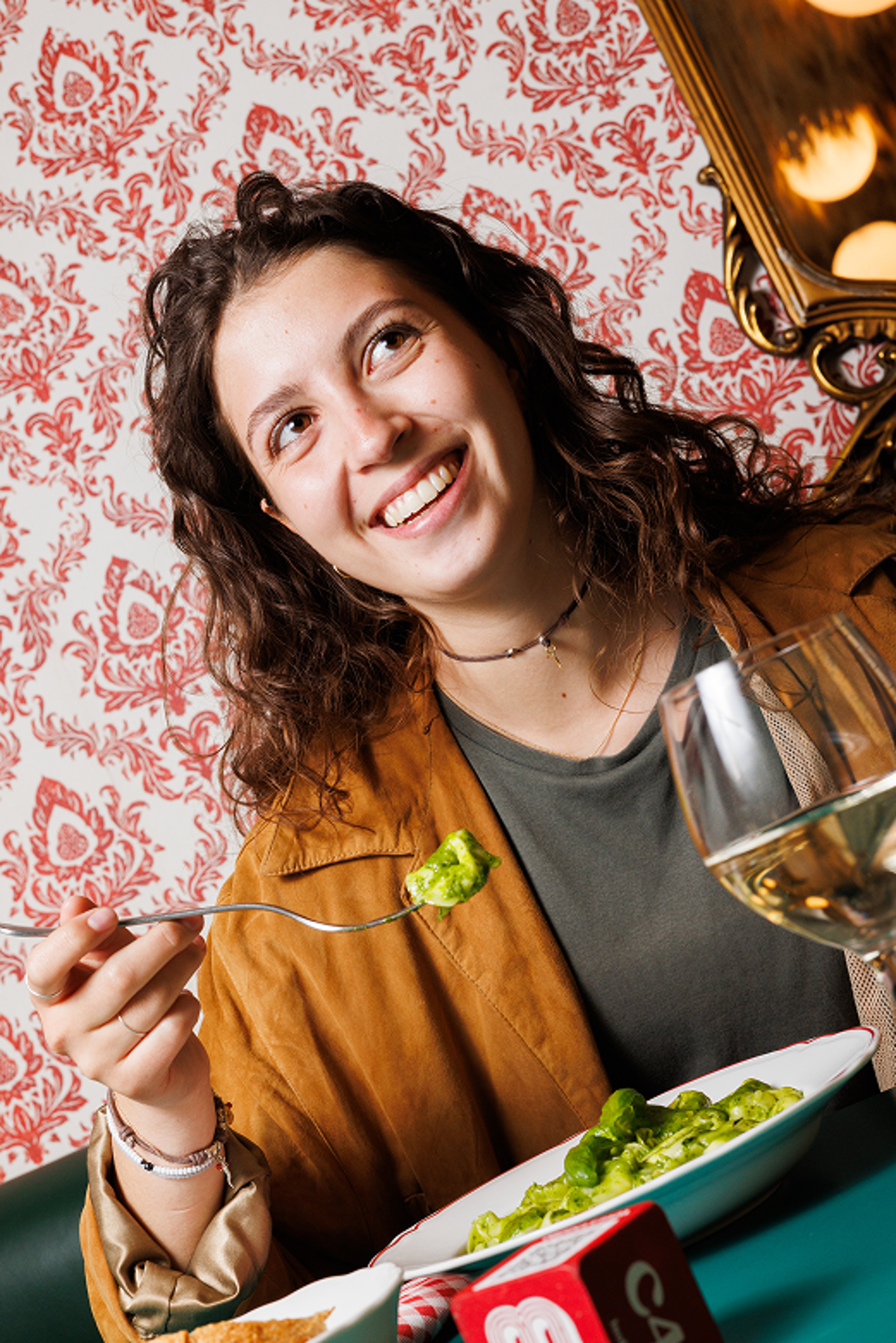 A smiling woman enjoys a plate of pesto pasta in a warmly lit restaurant with vintage wallpaper.