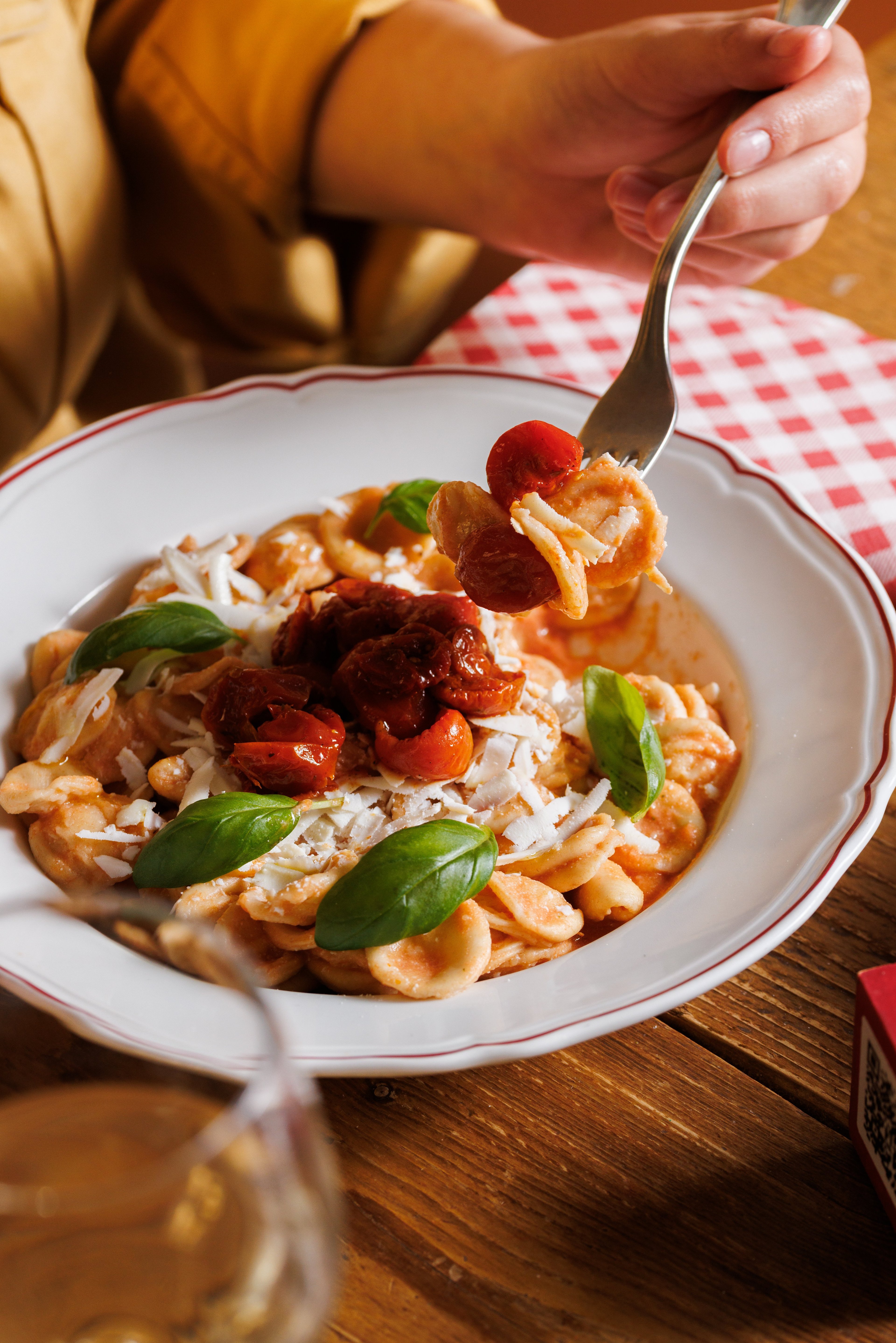 A person lifts a forkful of orecchiette pasta topped with sun-dried tomatoes, fresh basil, and grated cheese from a white plate.