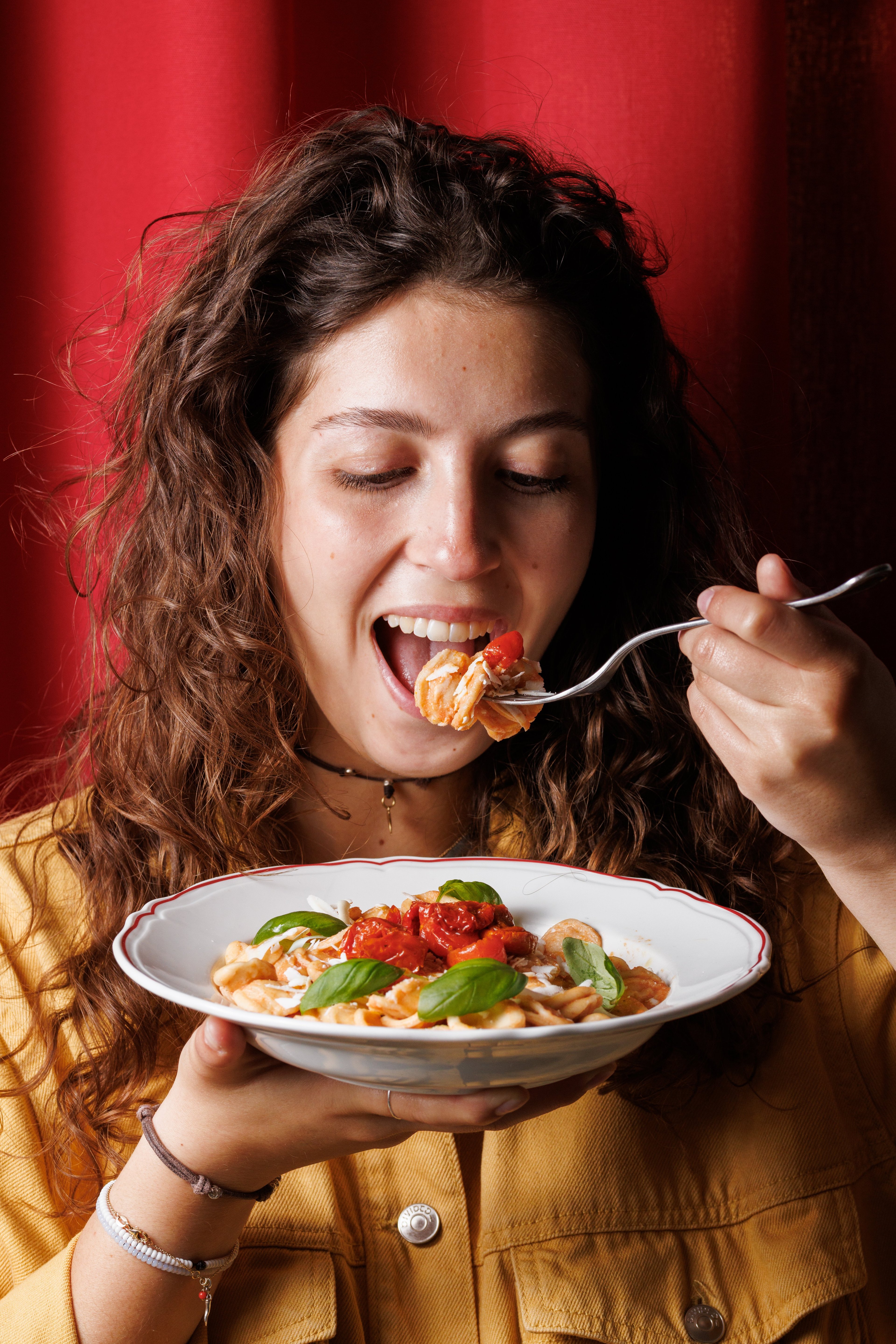 A young woman with curly hair enjoys a plate of fresh pasta topped with sun-dried tomatoes and basil in a warmly lit setting.