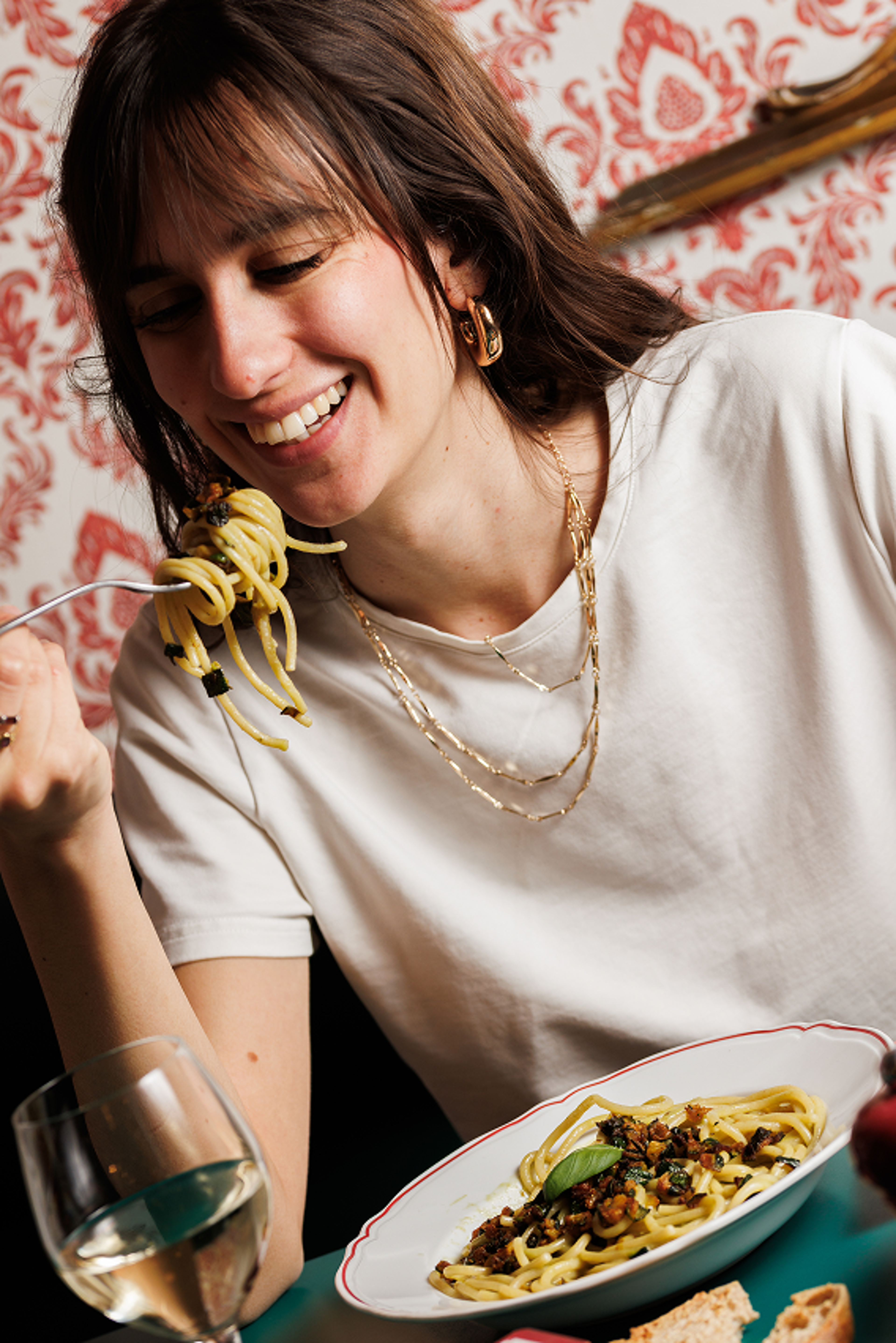 A woman smiles while eating a plate of pasta in a warmly lit restaurant with patterned wallpaper.