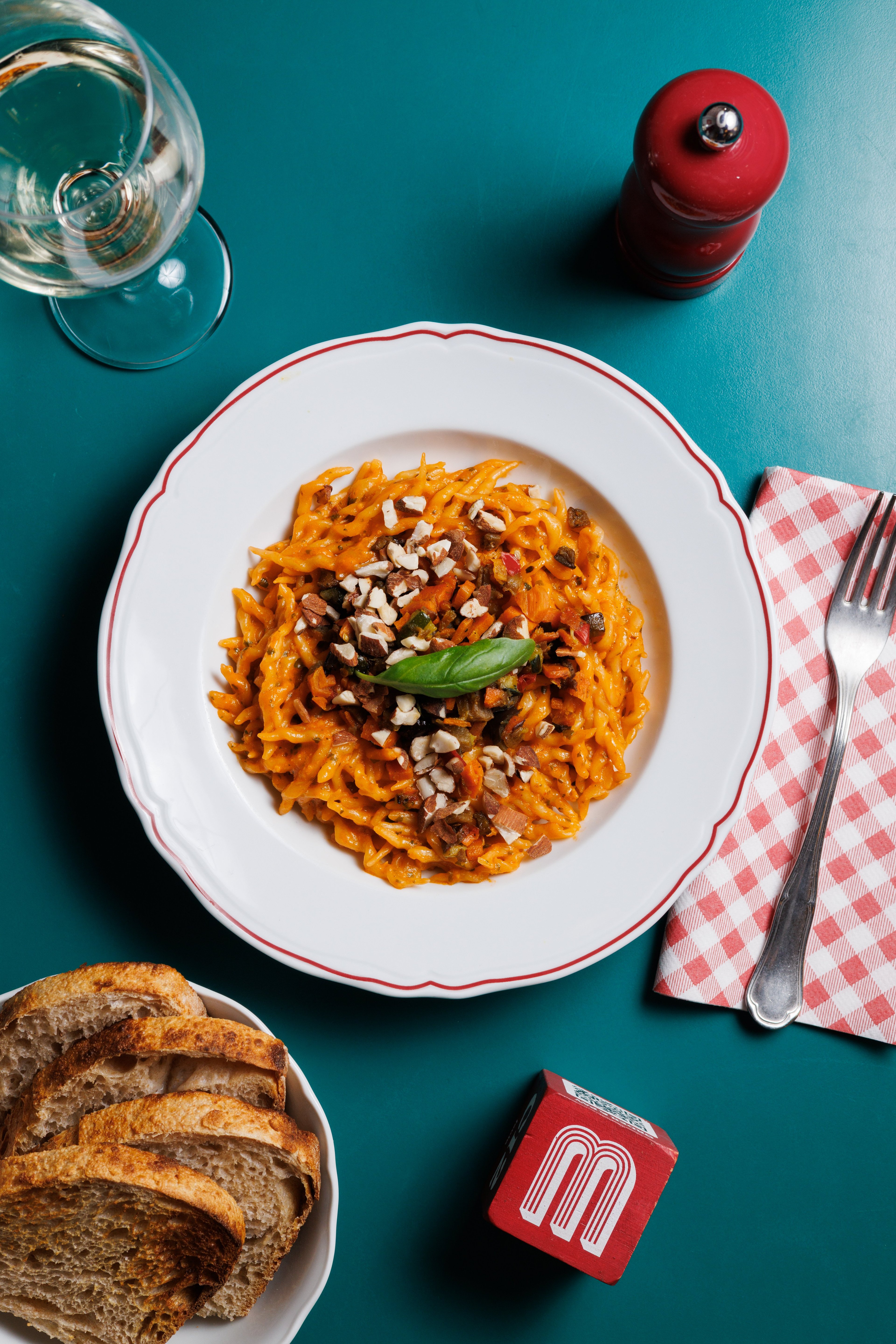 A top-down view of a plate of pasta with a creamy orange sauce, topped with chopped nuts and a fresh basil leaf, served on a teal table.