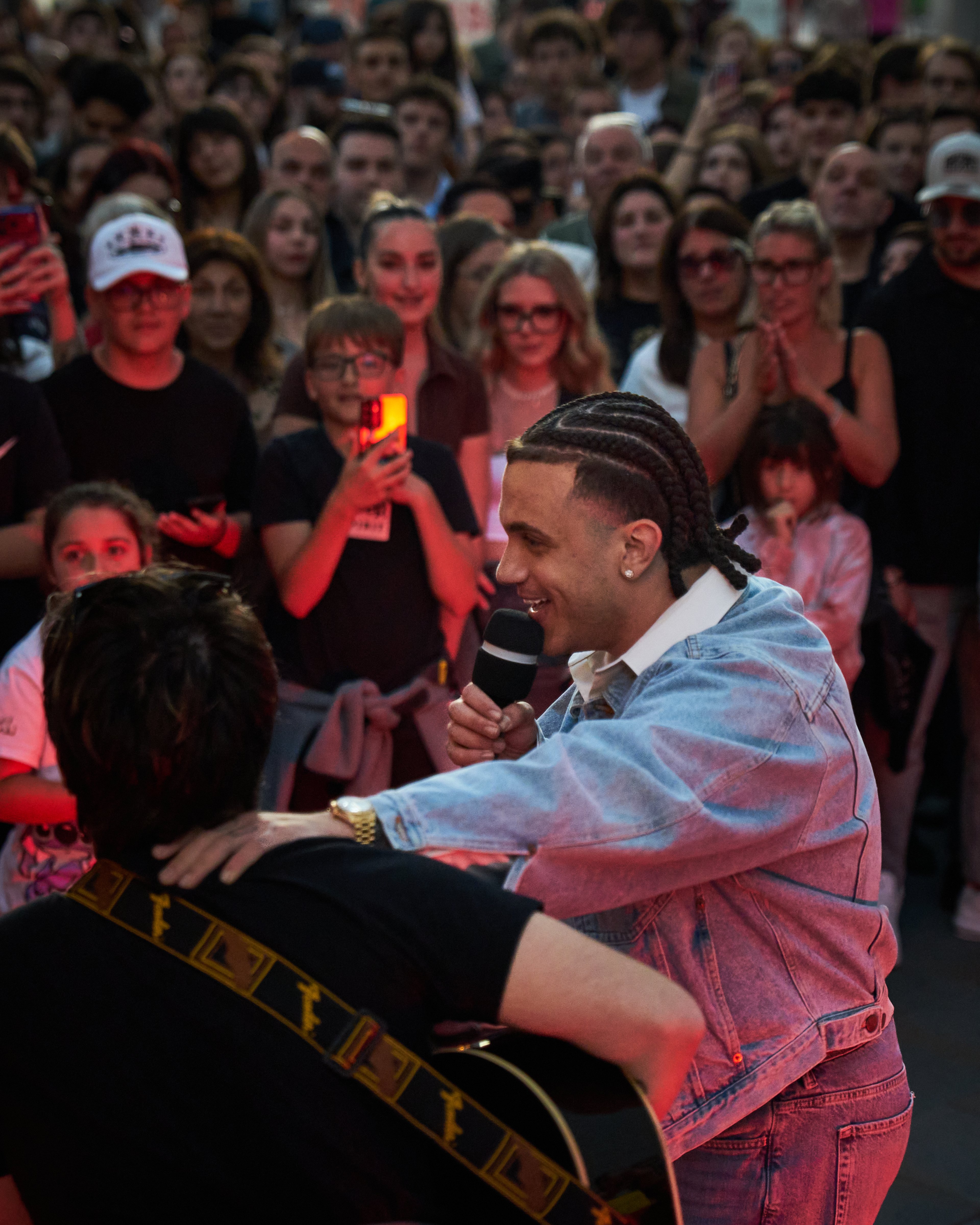 A performer with braided hair sings into a microphone while interacting with a guitarist in a crowded public space.