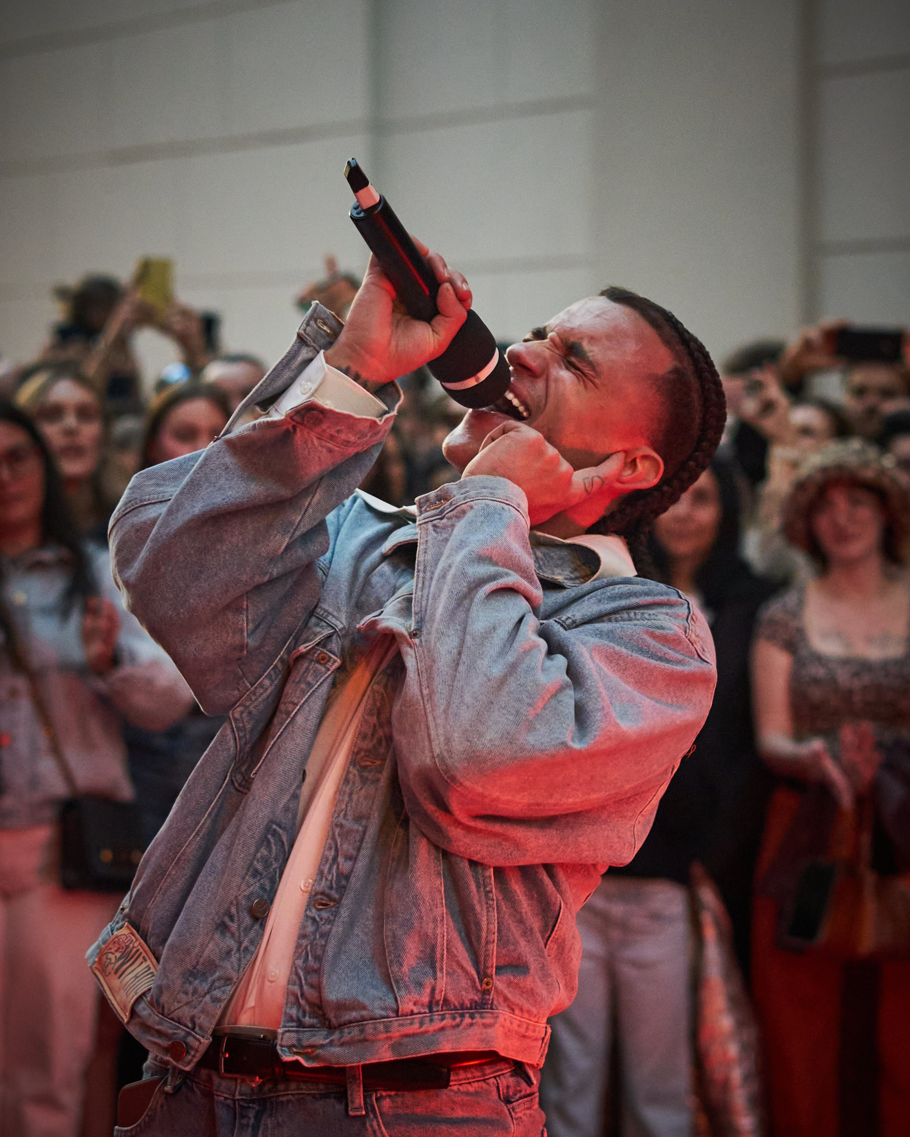 A performer sings passionately into a microphone while surrounded by an audience in a dimly lit, red-toned venue.