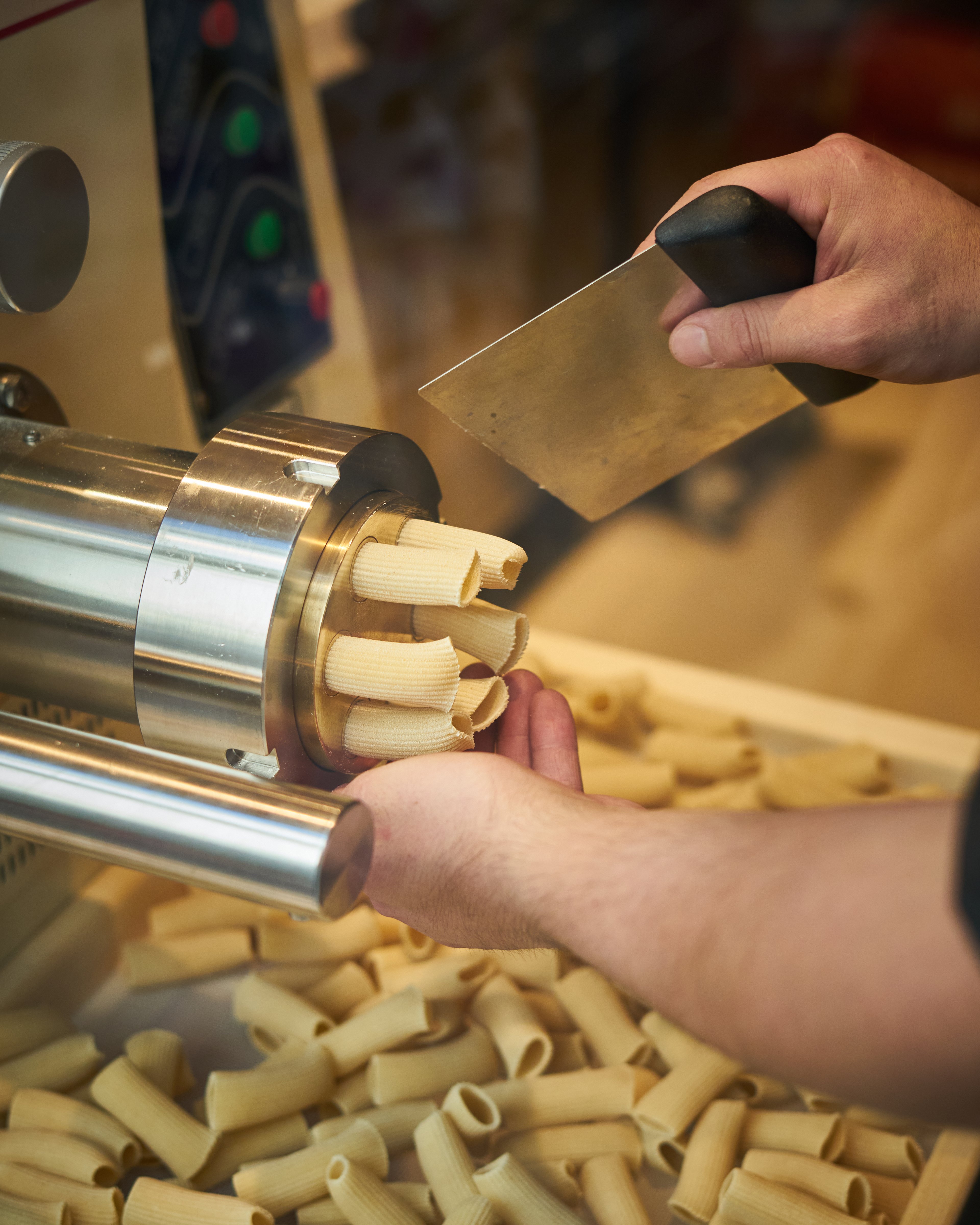 A chef uses a metal scraper to cut fresh rigatoni pasta as it extrudes from a commercial machine.
