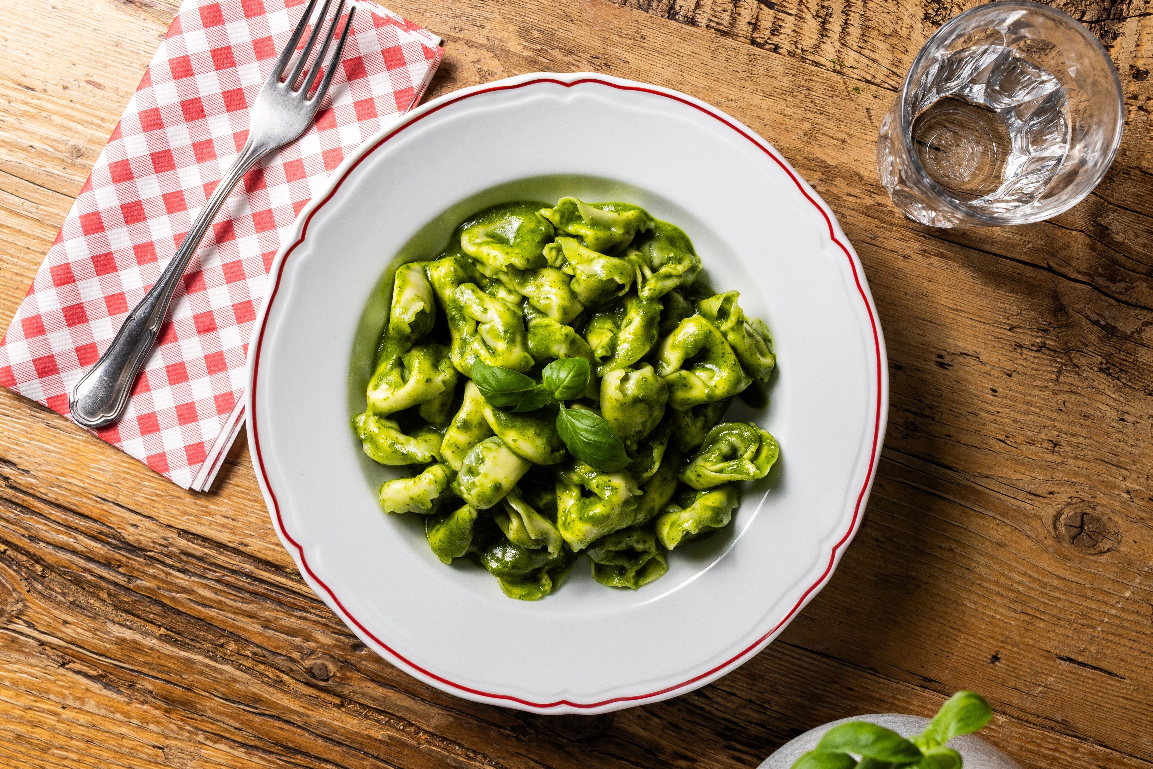 A top-down view of a plate of tortellini tossed in vibrant green pesto sauce, served on a rustic wooden table.