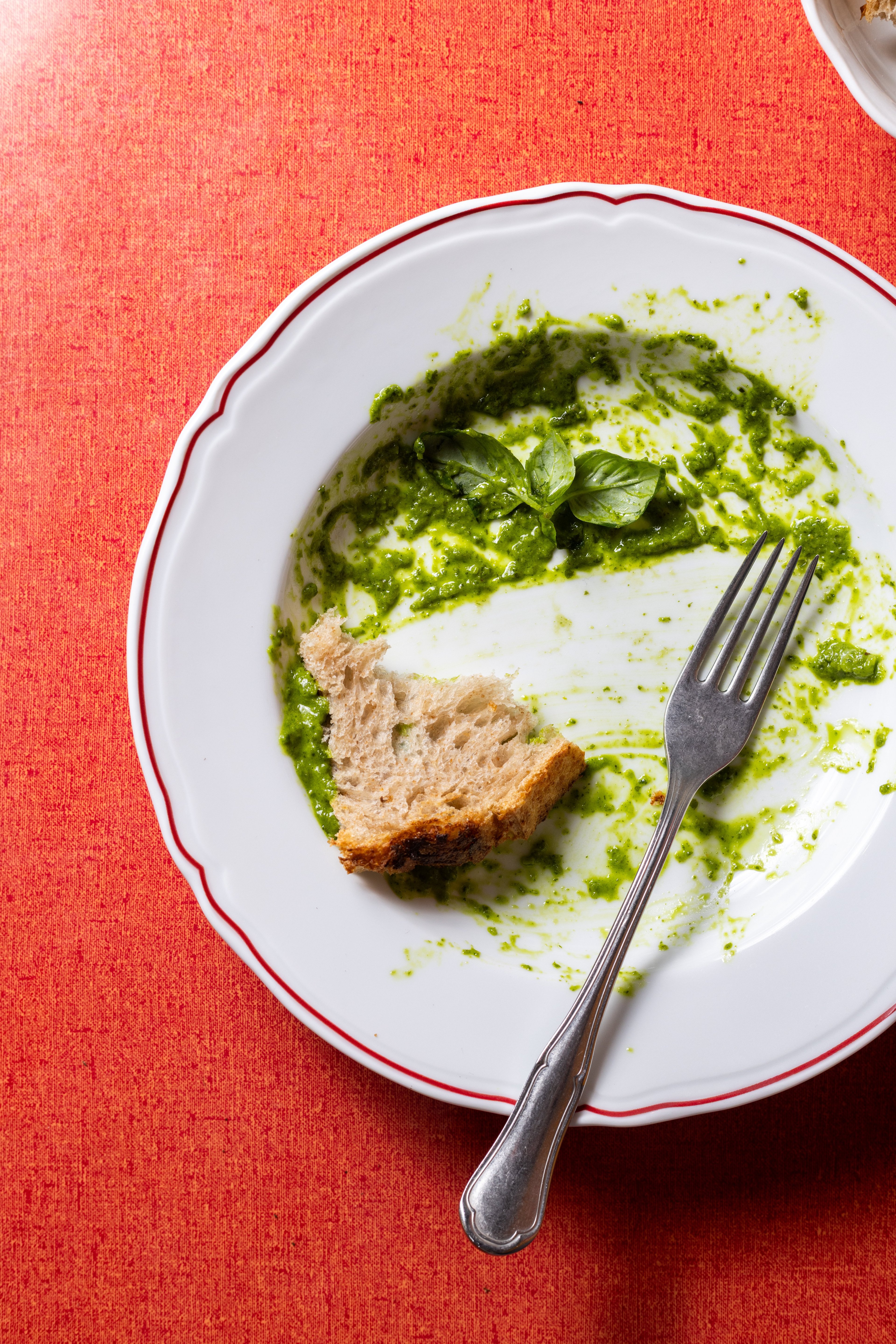 A partially eaten meal of pesto sauce and bread rests on a white plate with a fork against a vibrant orange tablecloth.