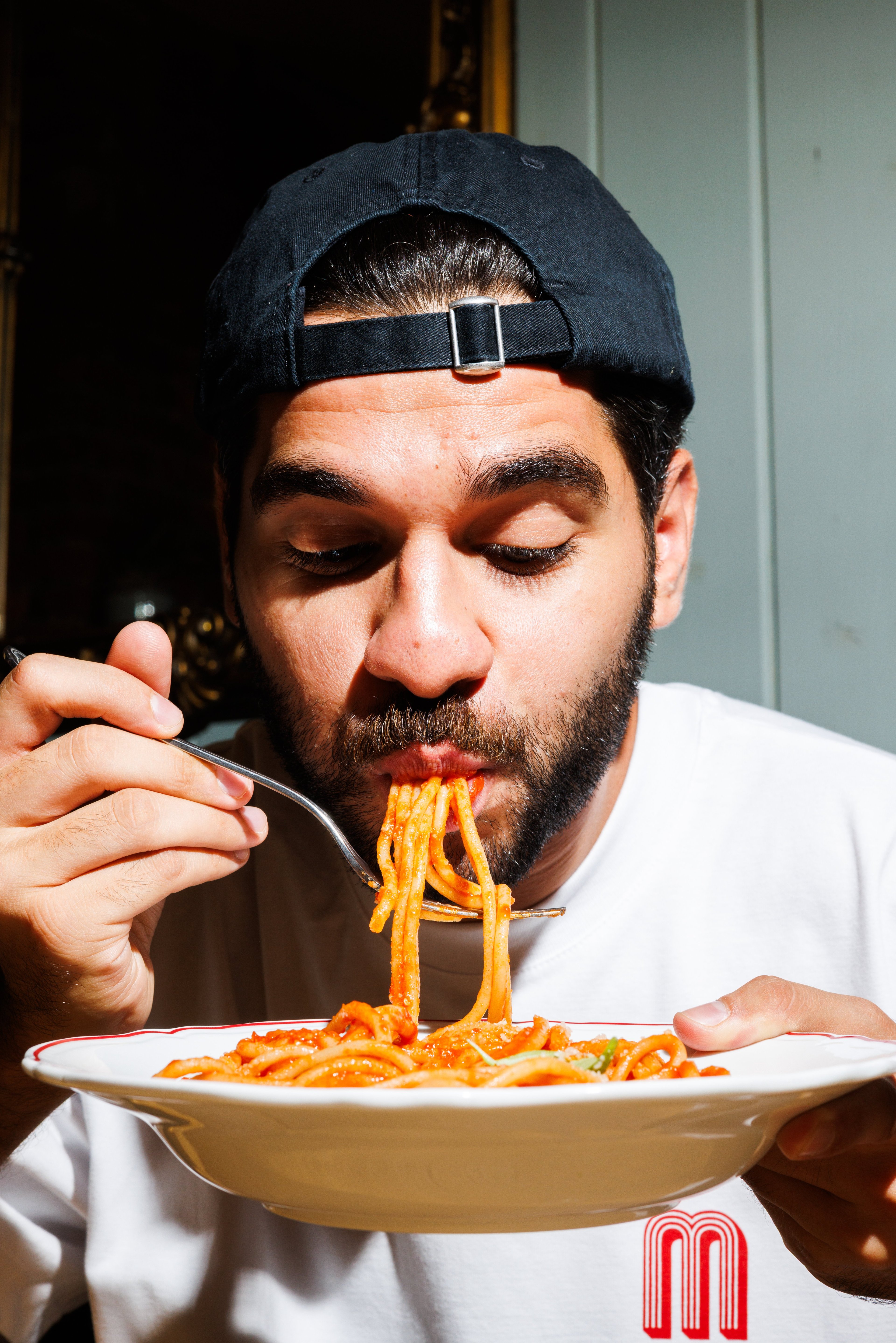 A man wearing a baseball cap enjoys a plate of spaghetti with tomato sauce in a brightly lit restaurant setting.