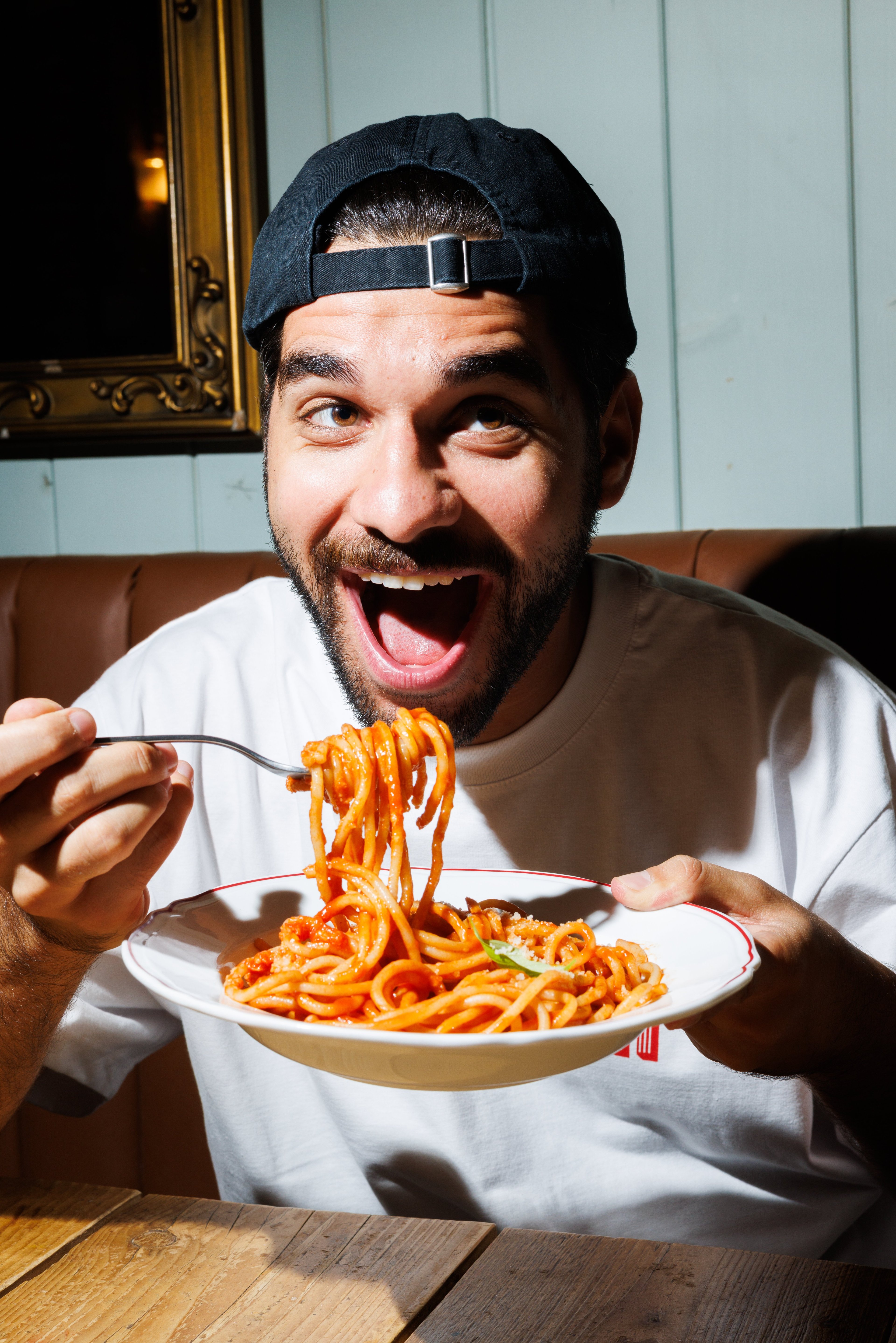 A man with a beard and a baseball cap excitedly prepares to eat a plate of spaghetti with tomato sauce in a dimly lit restaurant.