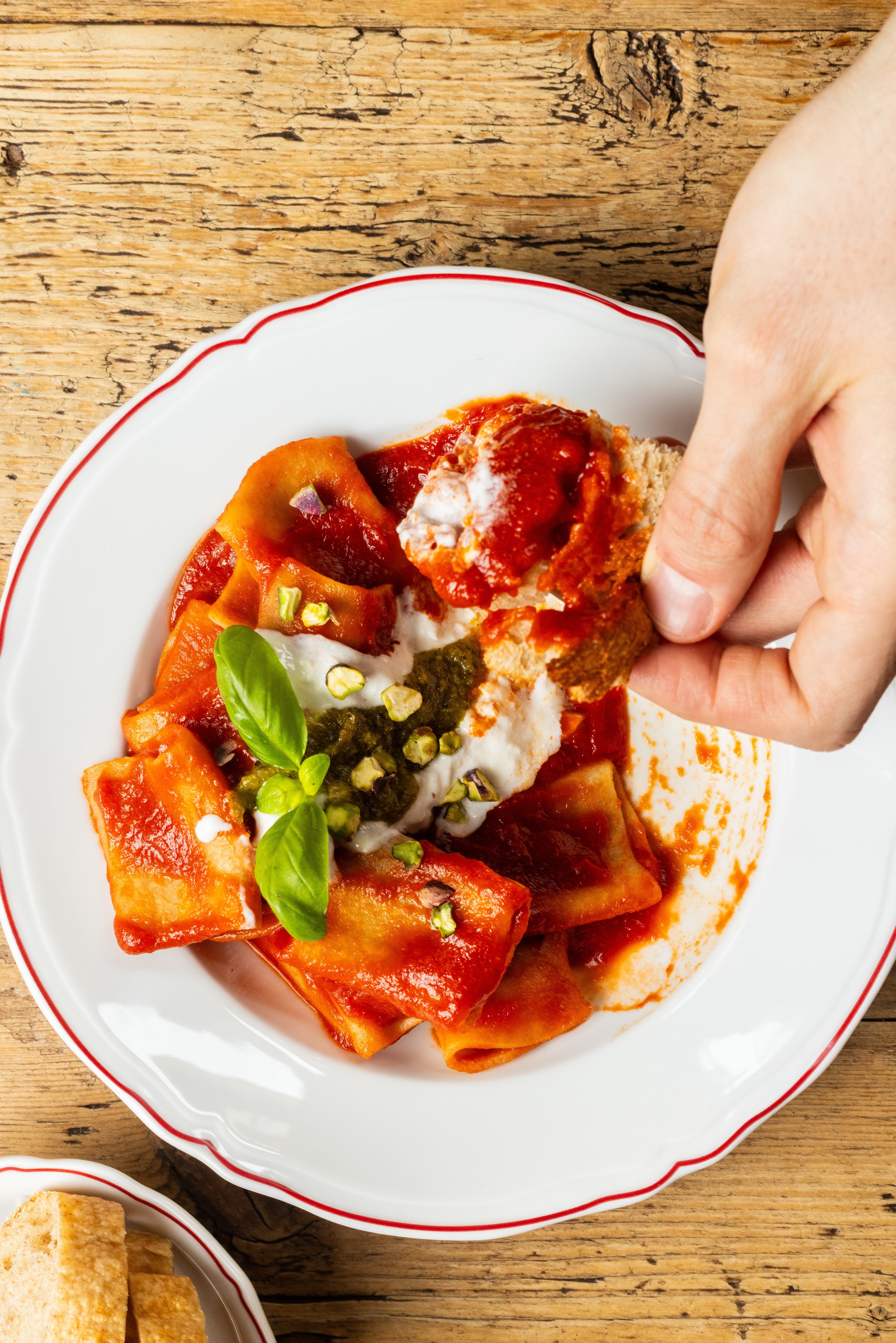 A hand dips a piece of crusty bread into a plate of pasta topped with tomato sauce, creamy cheese, pesto, and pistachios.