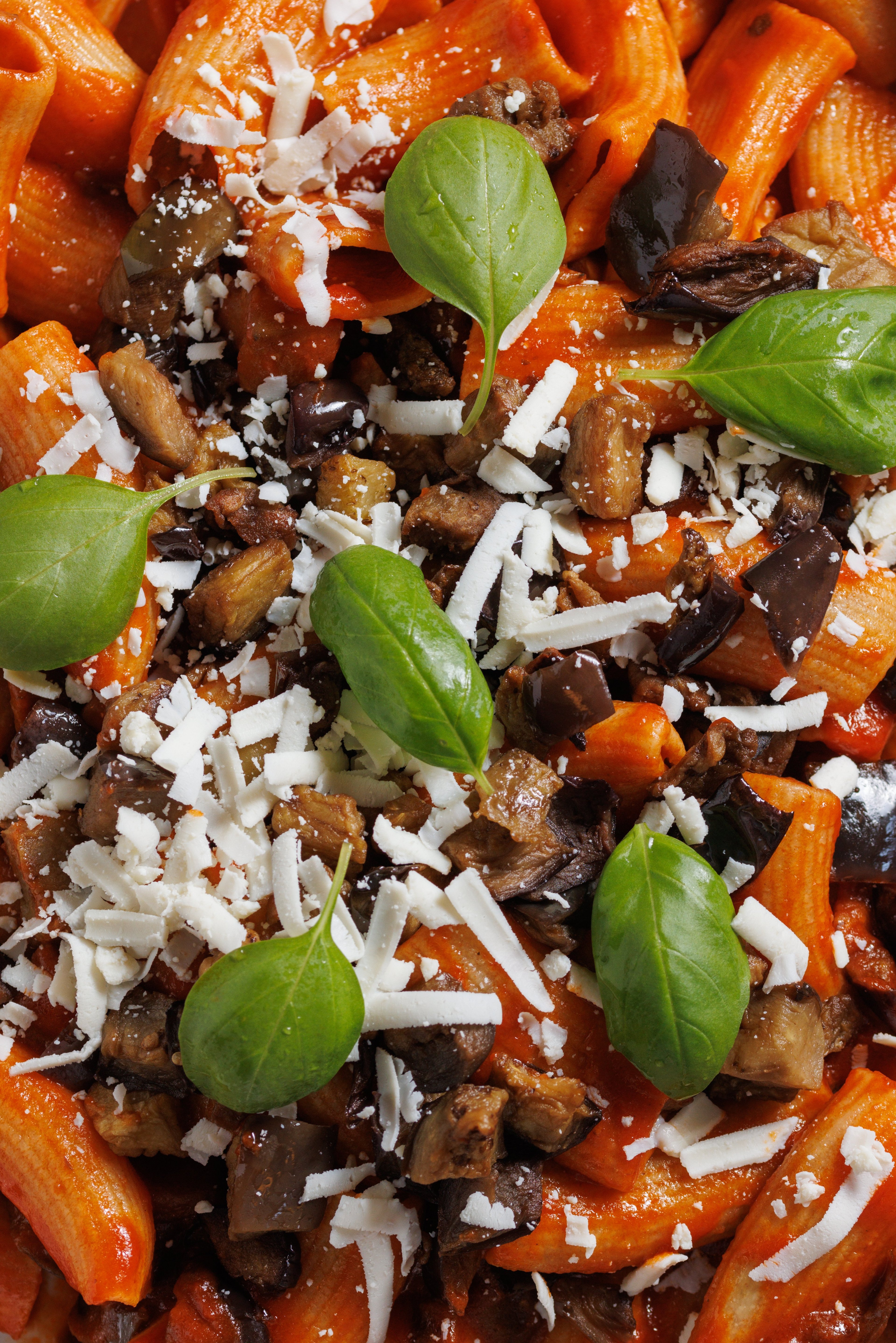 A close-up overhead shot of rigatoni pasta tossed in tomato sauce with roasted eggplant, fresh basil, and grated cheese.