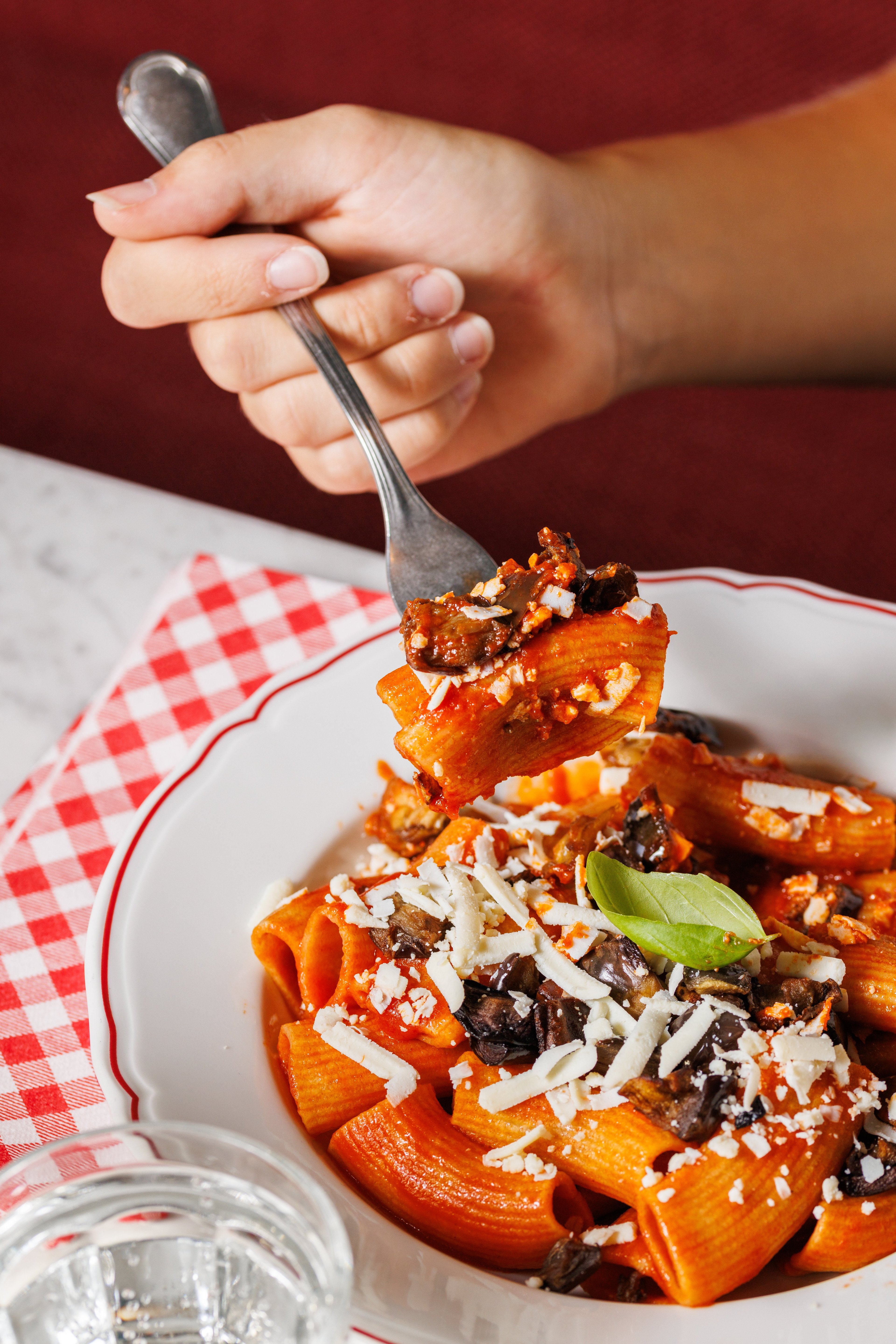 A hand holds a fork with a piece of rigatoni pasta topped with tomato sauce, eggplant, and grated cheese.