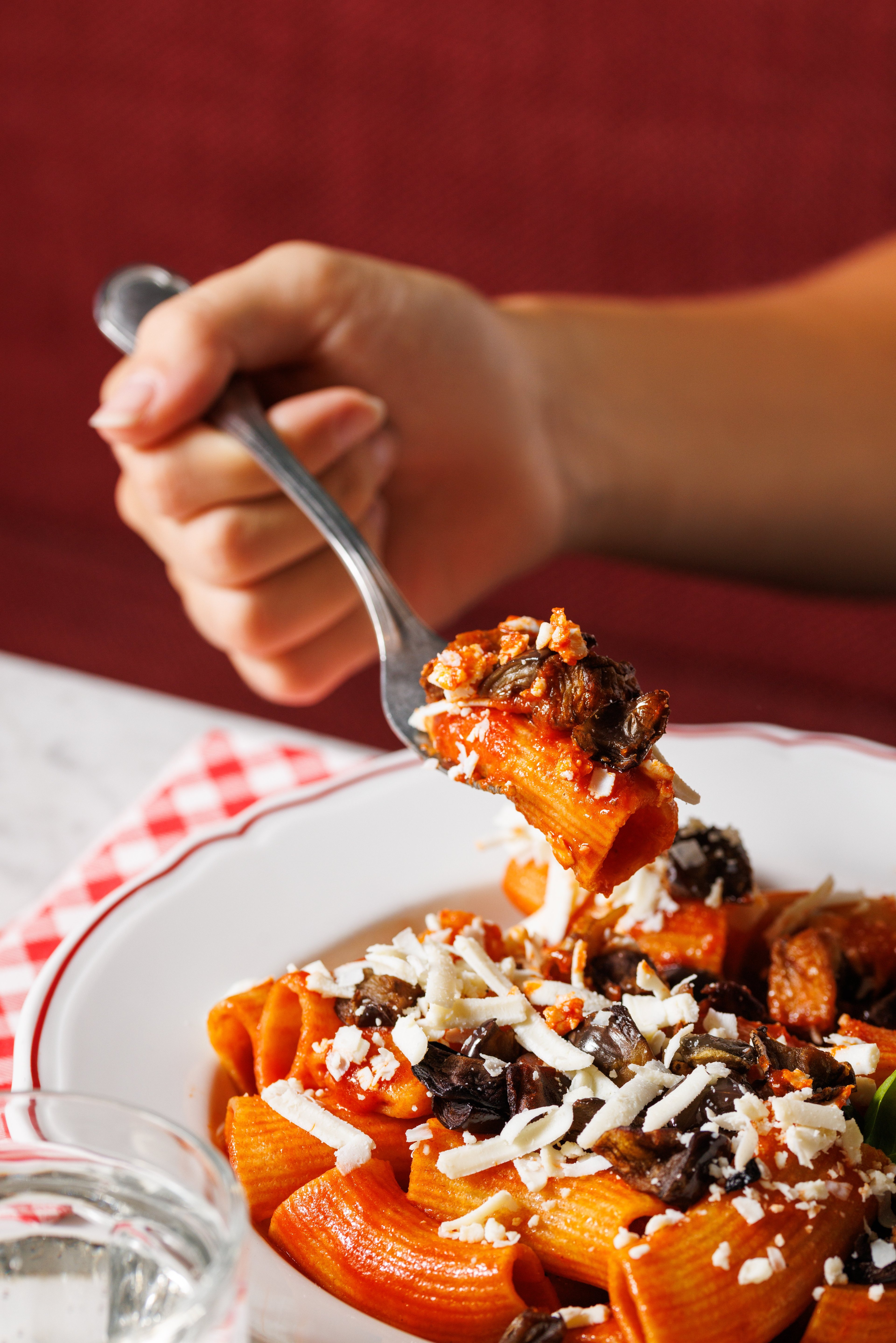 A hand holds a fork lifting a piece of rigatoni pasta topped with tomato sauce, eggplant, and grated cheese.