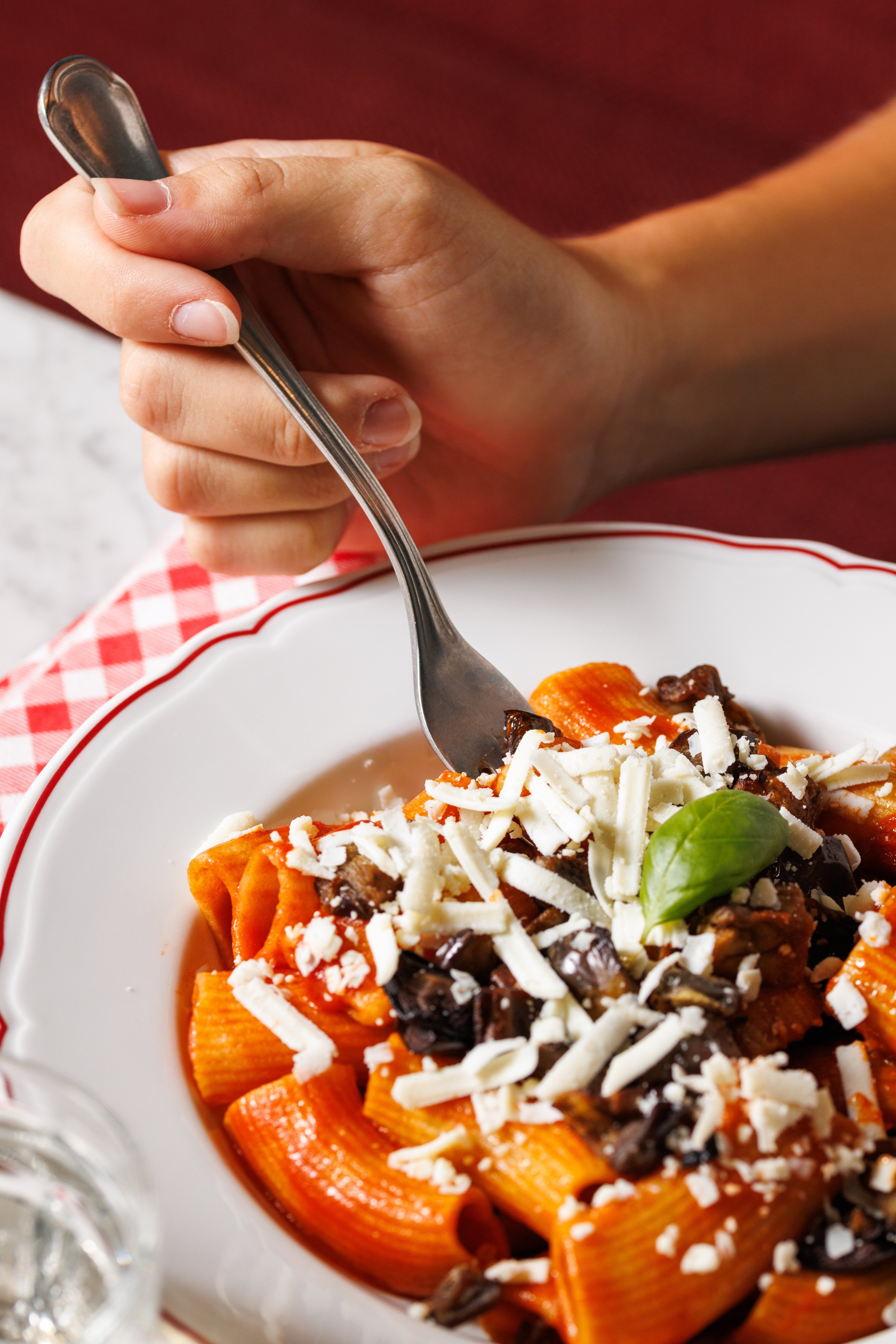 A close-up view of a hand using a fork to eat a plate of rigatoni pasta topped with tomato sauce, eggplant, and grated cheese.