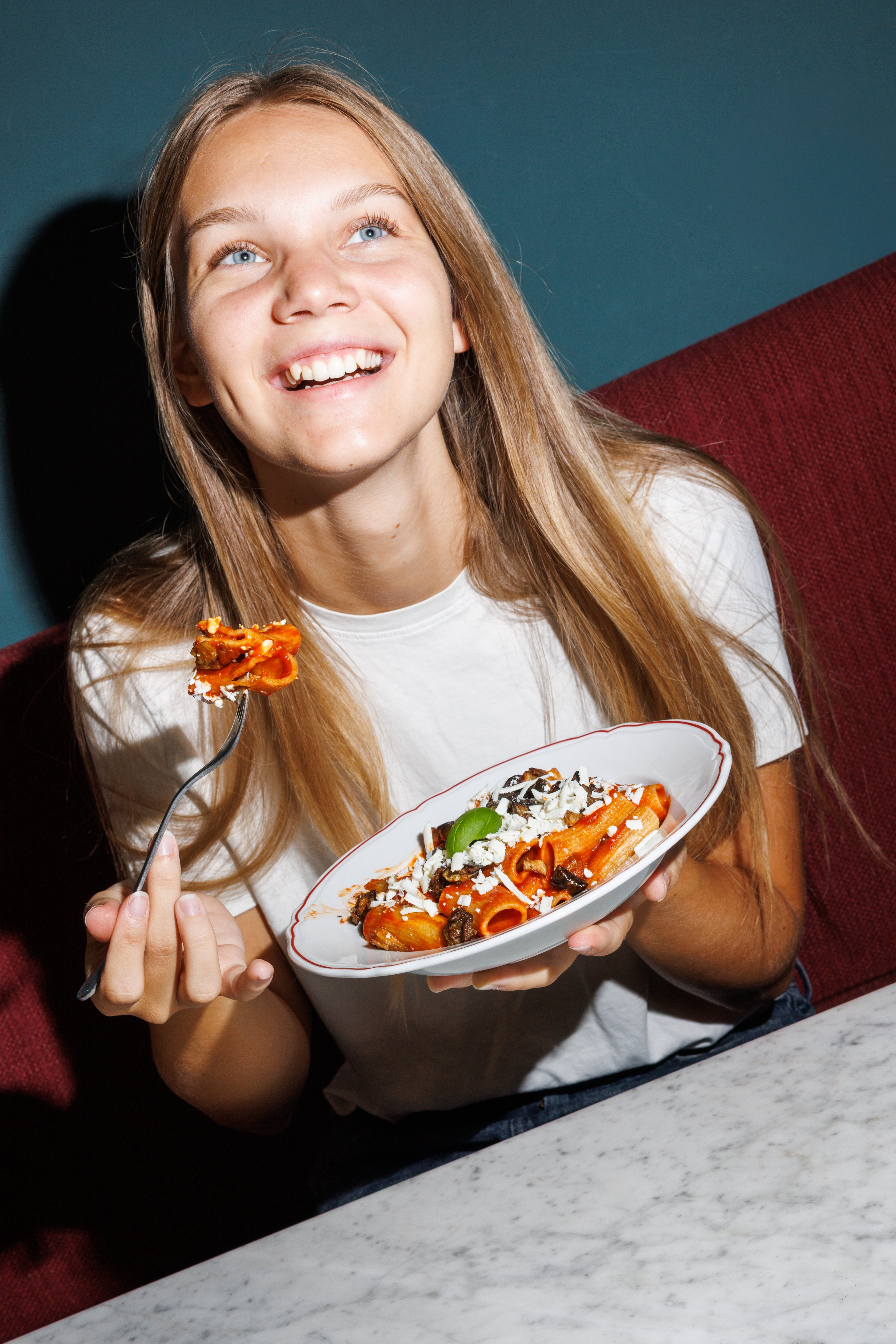 A smiling young woman enjoys a plate of rigatoni pasta topped with tomato sauce, mushrooms, and cheese in a cozy restaurant setting.