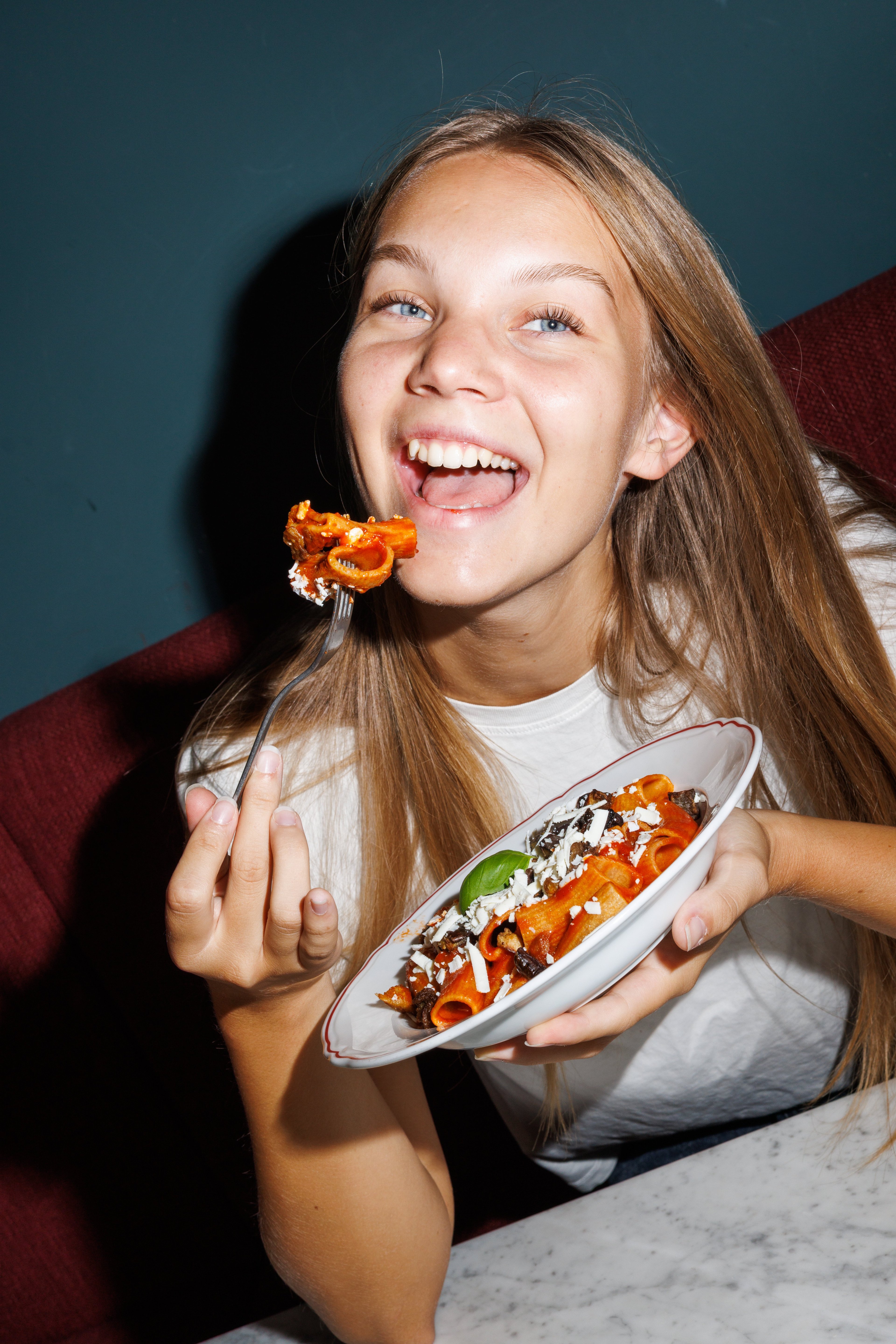 A smiling young woman enjoys a plate of rigatoni pasta with tomato sauce and cheese in a dimly lit restaurant.
