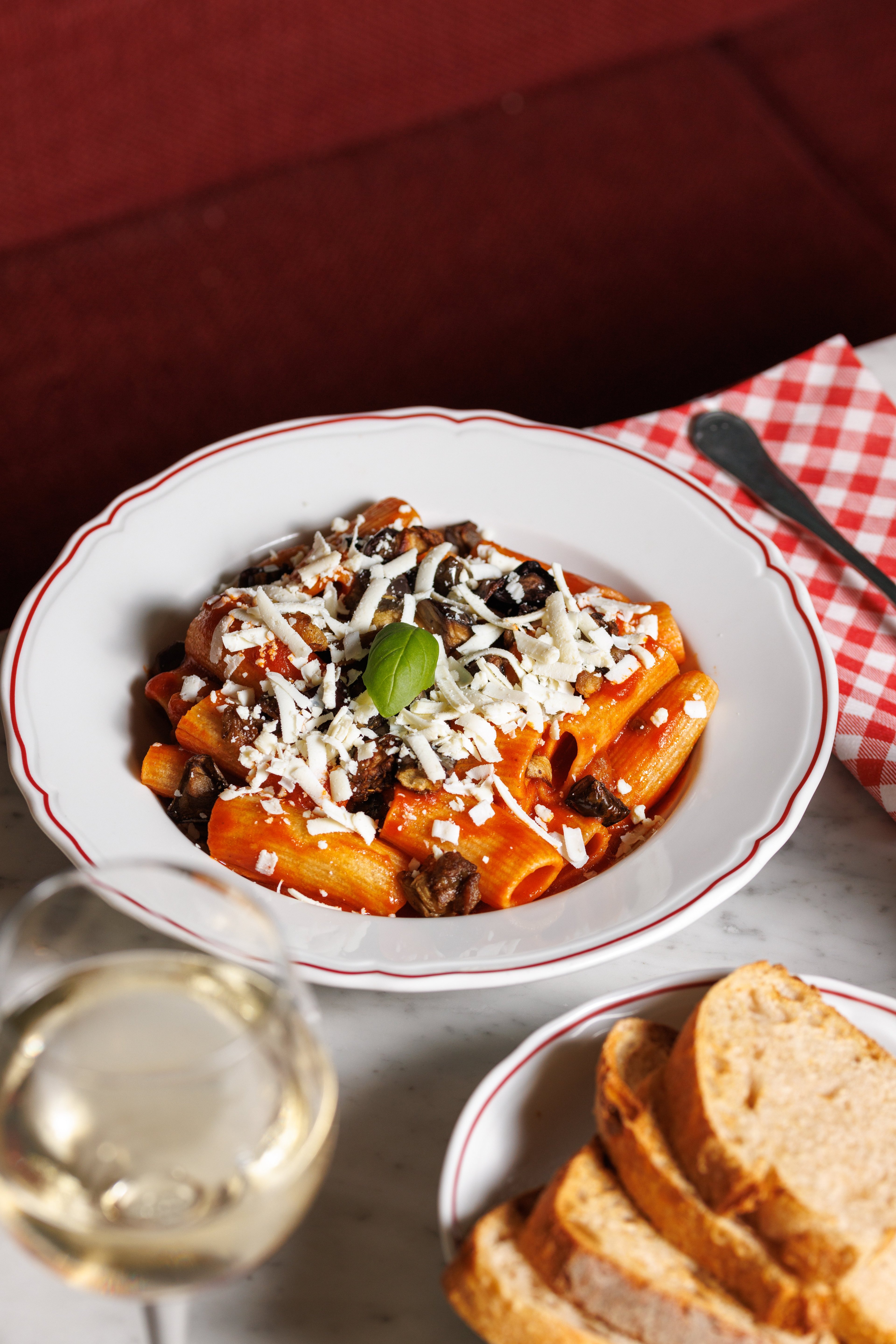 A plate of rigatoni pasta with tomato sauce, eggplant, and grated cheese is served on a marble table alongside bread and white wine.