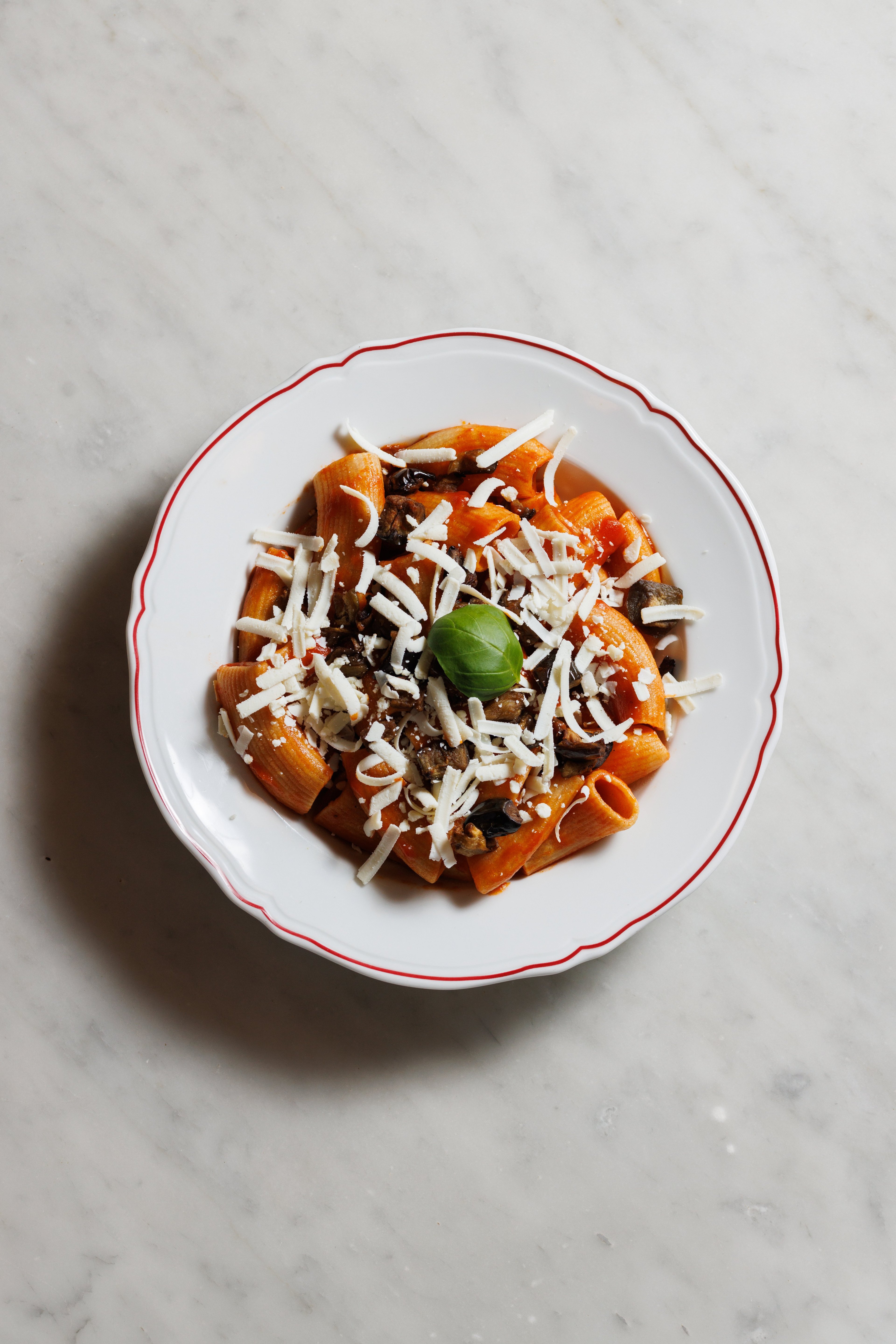 A plate of rigatoni pasta with tomato sauce, eggplant, and shredded cheese topped with a fresh basil leaf.