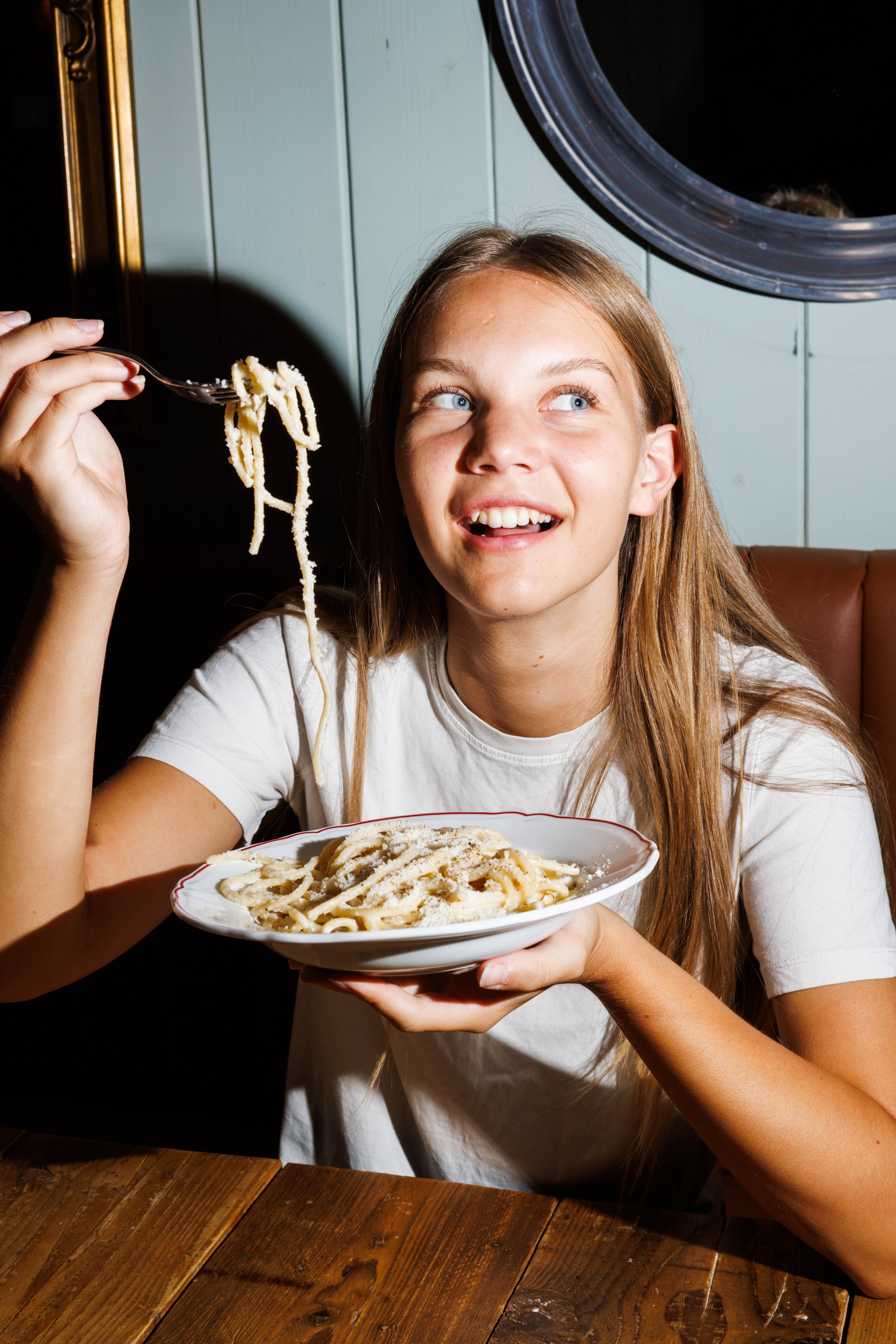 A young woman smiles while holding a forkful of pasta over a plate of cacio e pepe in a dimly lit restaurant.
