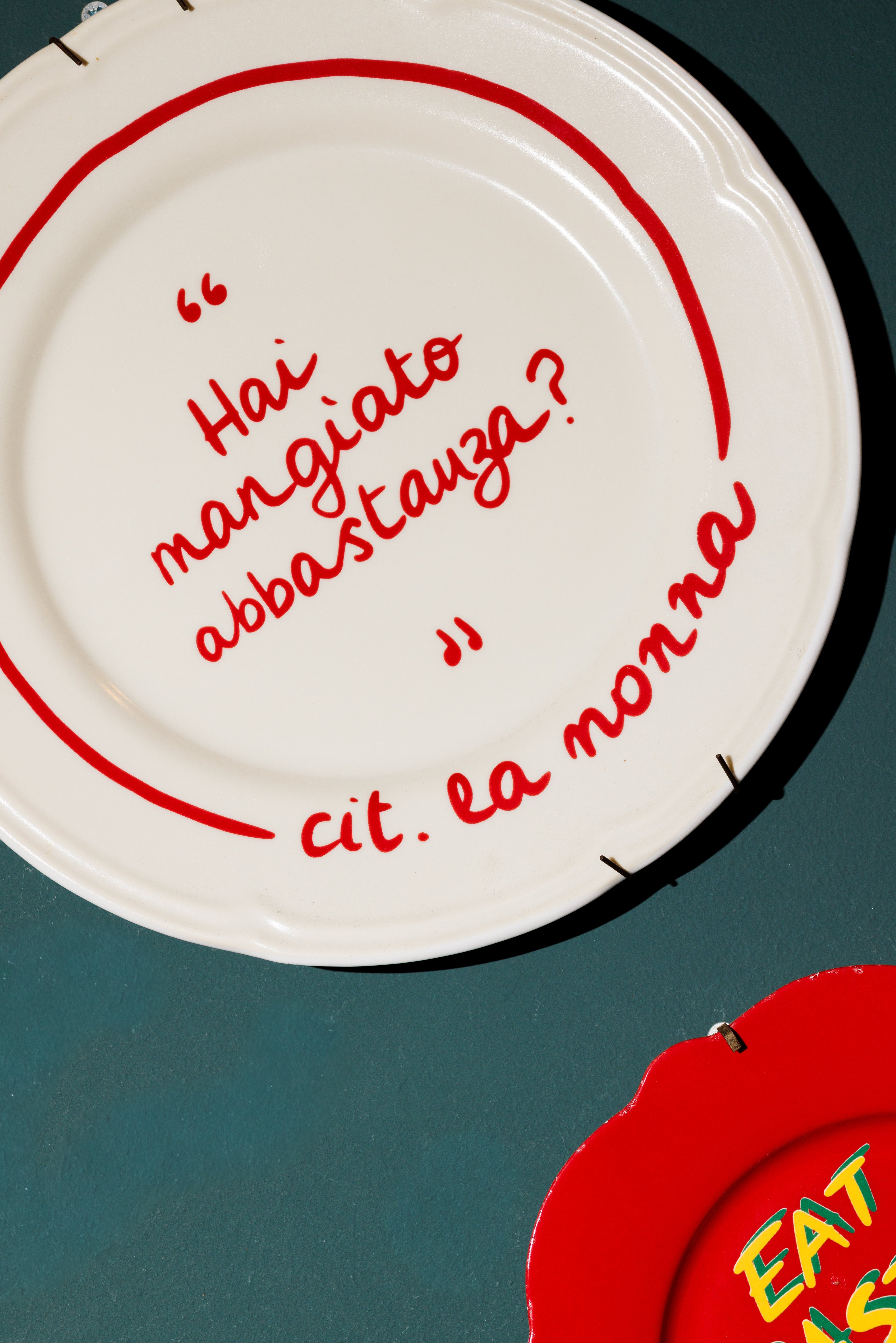 A decorative wall plate features a playful Italian quote about eating, displayed against a dark teal background.