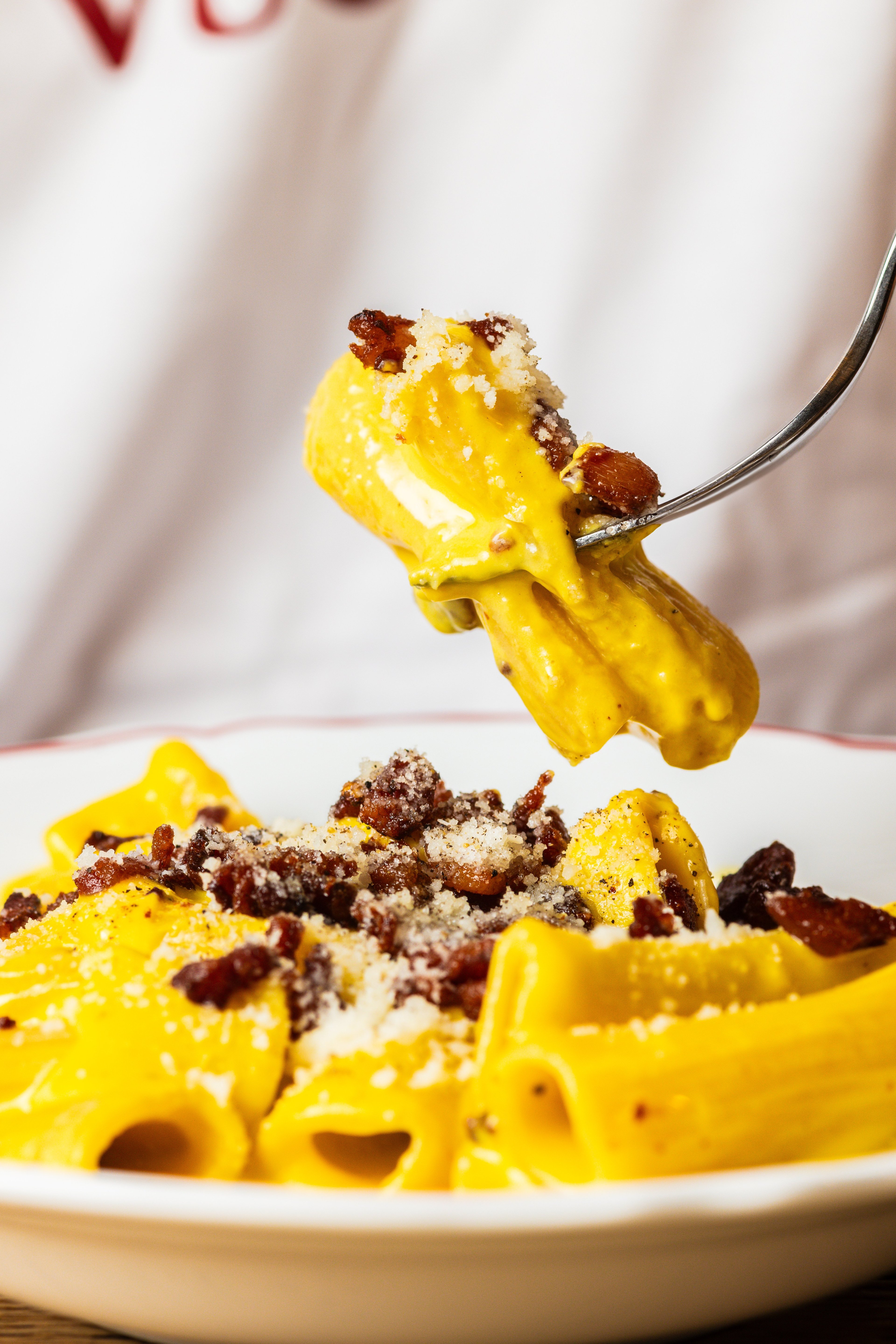 A close-up shot of a fork lifting a piece of creamy yellow pasta topped with crispy bacon and grated cheese.