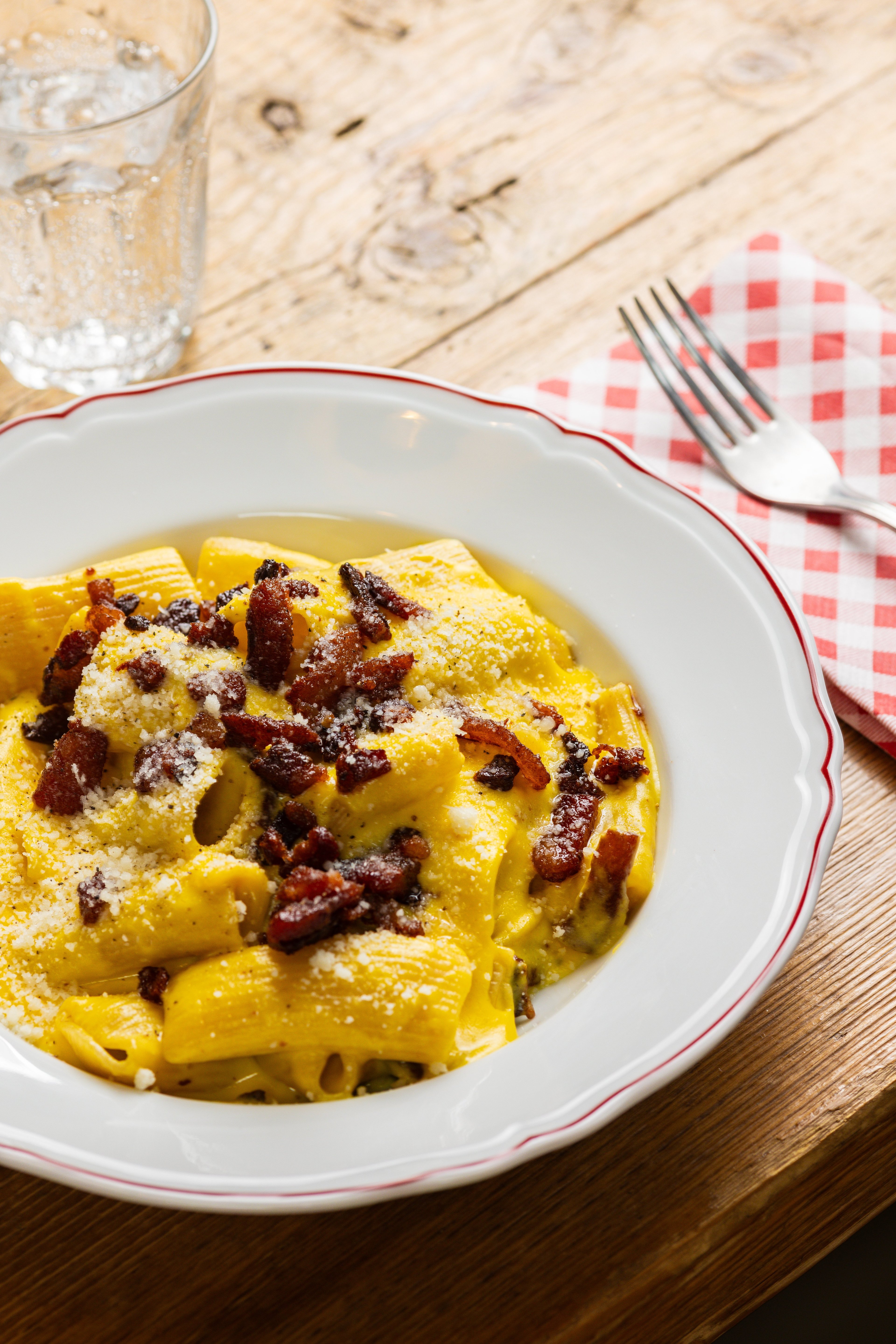 A plate of creamy pasta topped with crispy bacon and grated cheese sits on a rustic wooden table next to a glass of water and a checkered napkin.