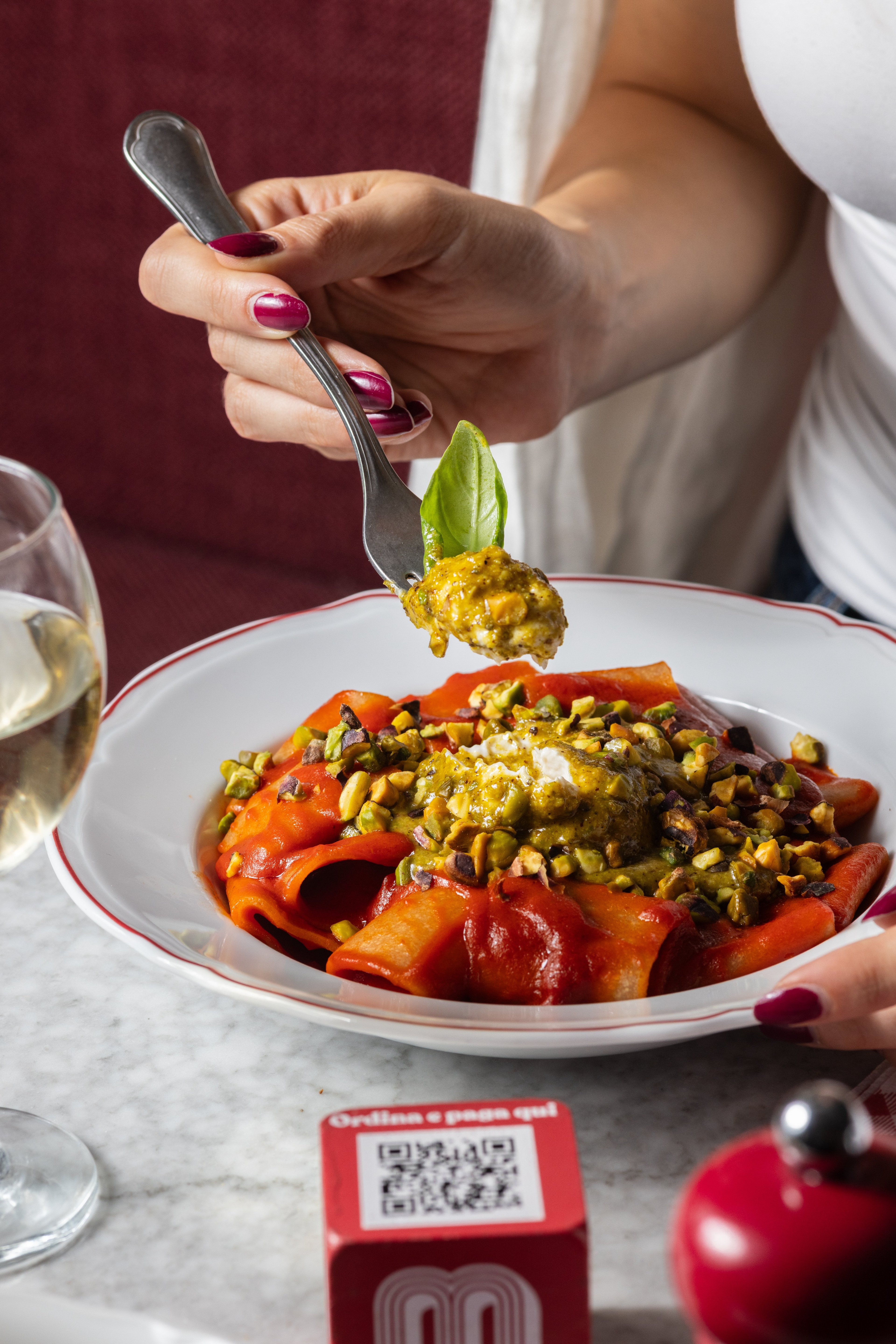 A person uses a fork to lift a bite of pasta topped with pesto, pistachios, and a basil leaf from a plate of red-sauced pasta.