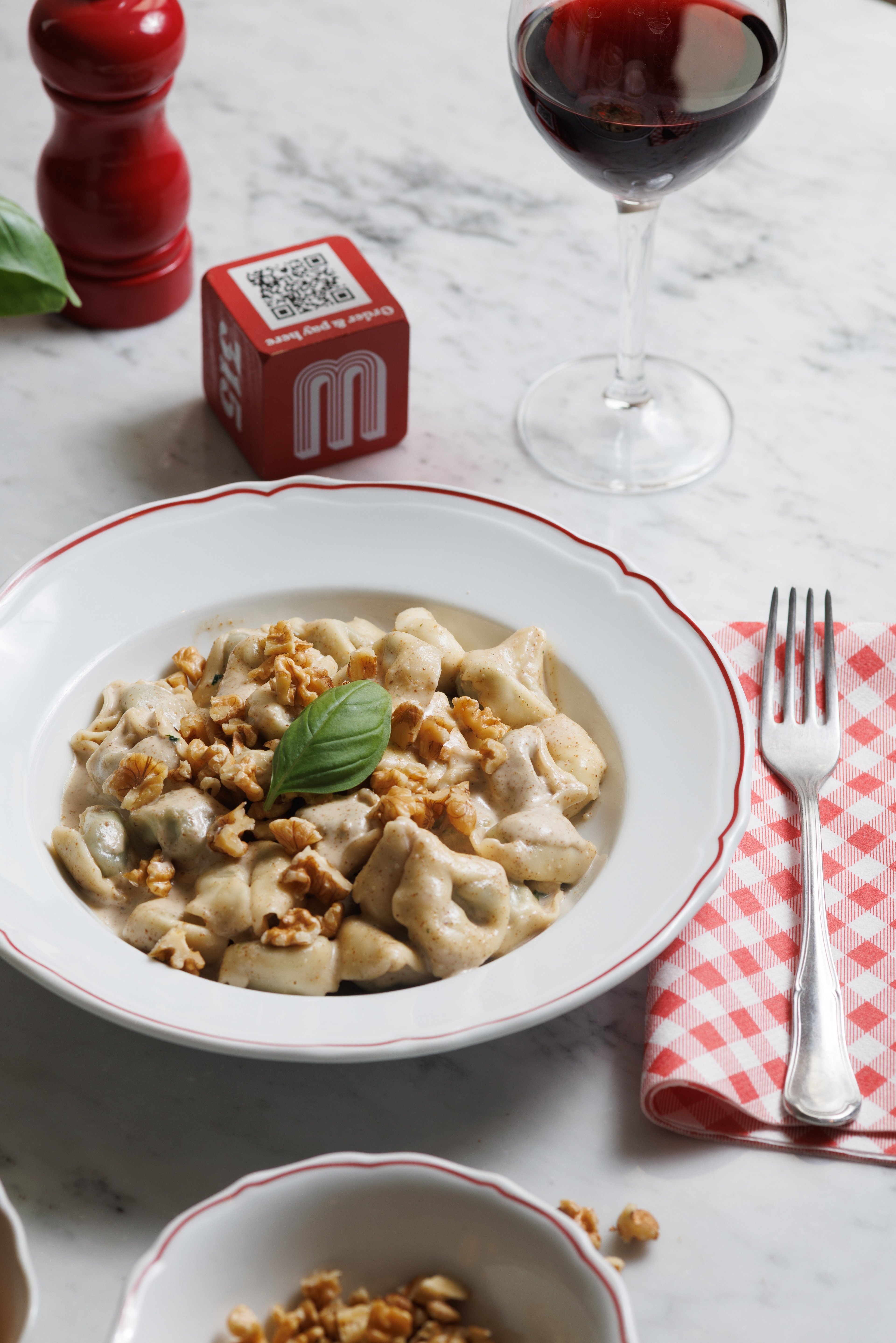 A plate of creamy walnut tortellini is served on a marble table alongside a glass of red wine and a checkered napkin.